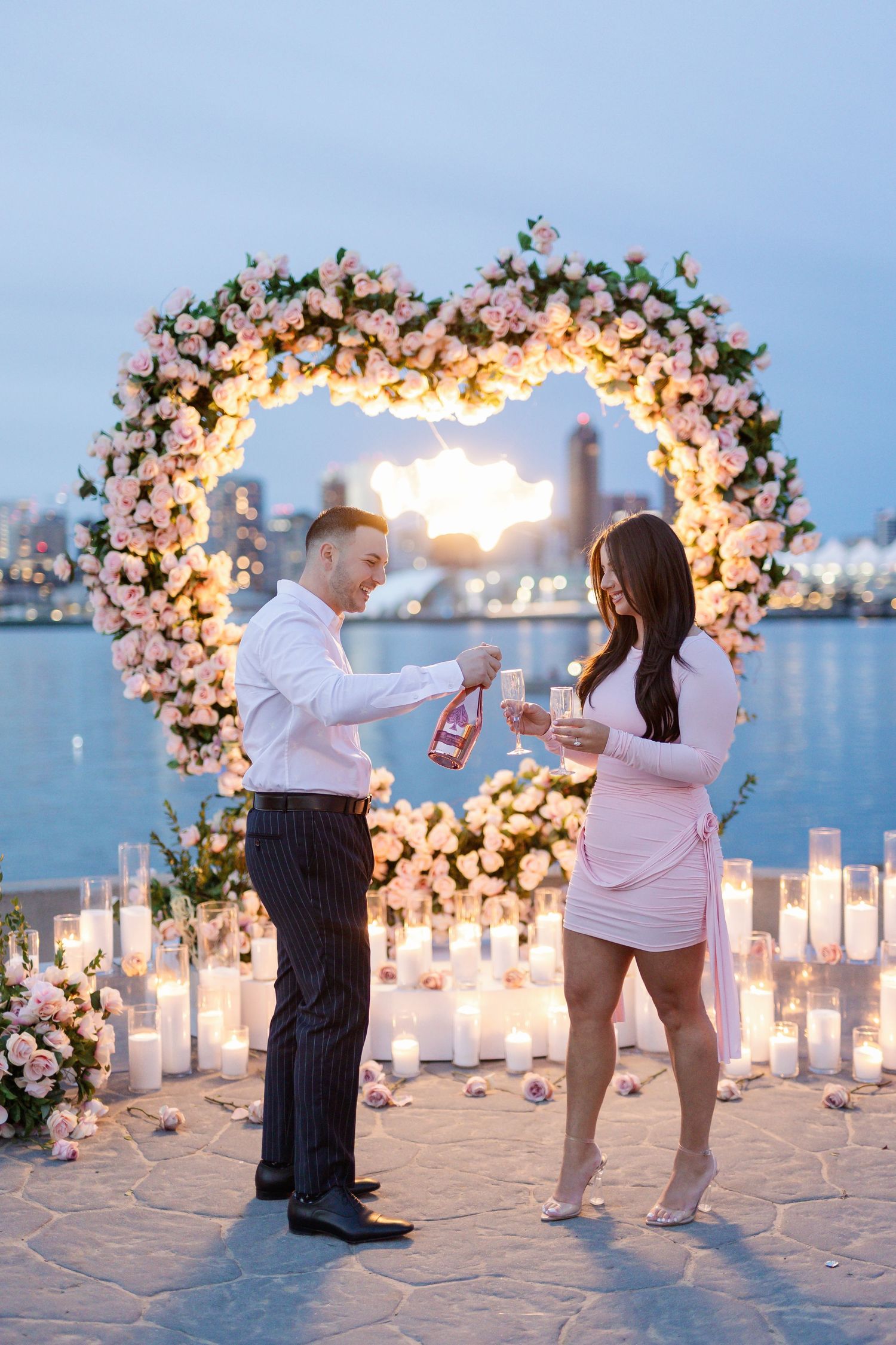 Couple celebrating proposal moment under floral arch with candles and city lights.