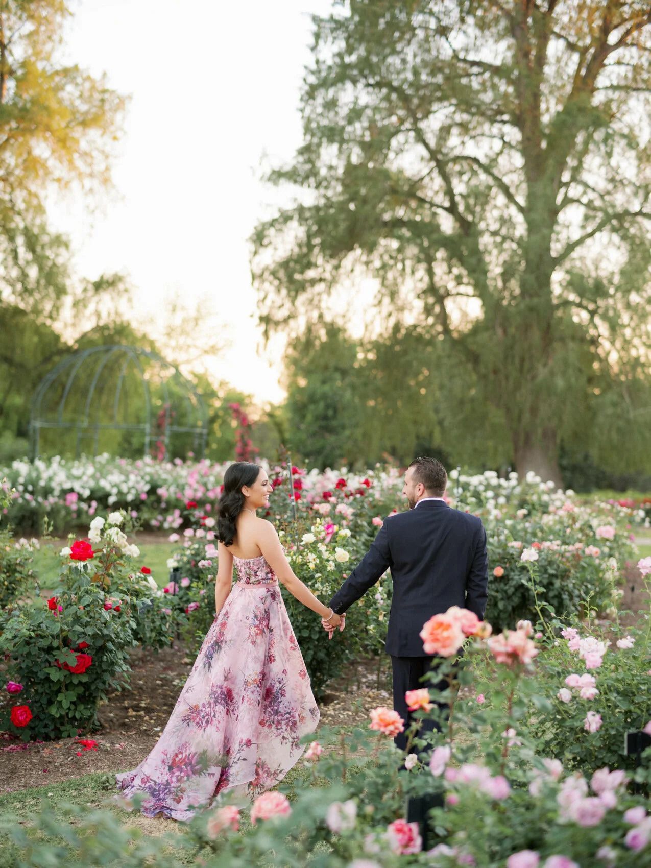 Couple walking through a blooming rose garden at sunset in formal attire.