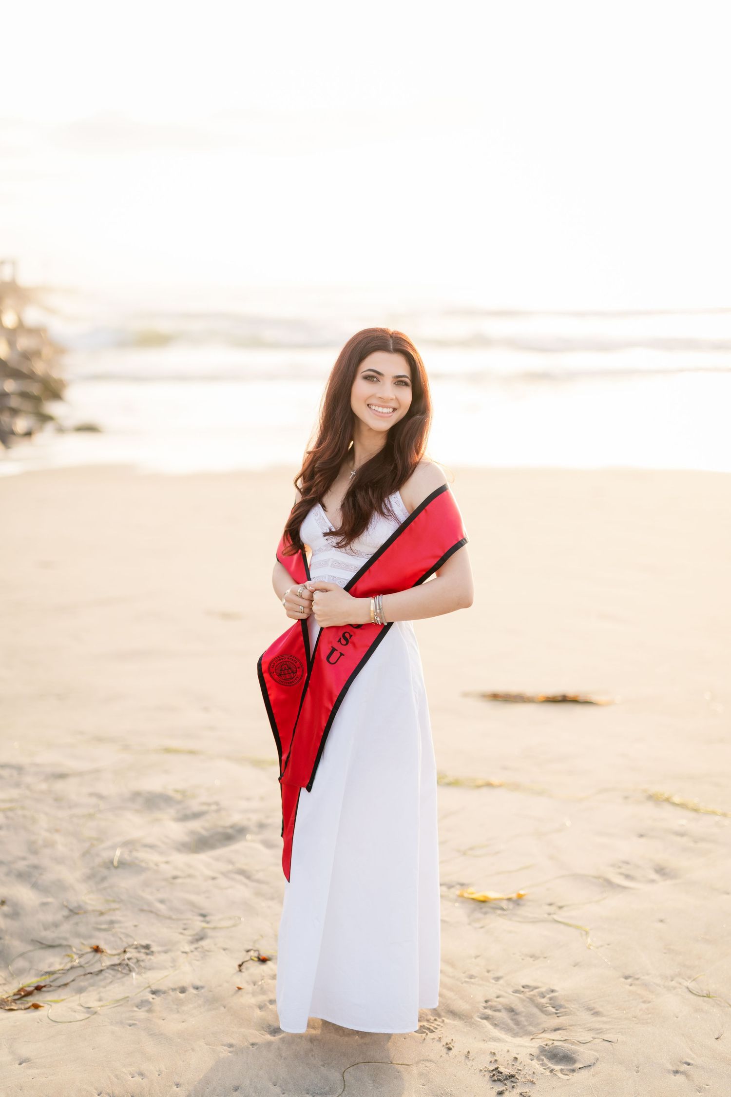A graduate in white dress with red sash poses on a sandy beach during golden hour.