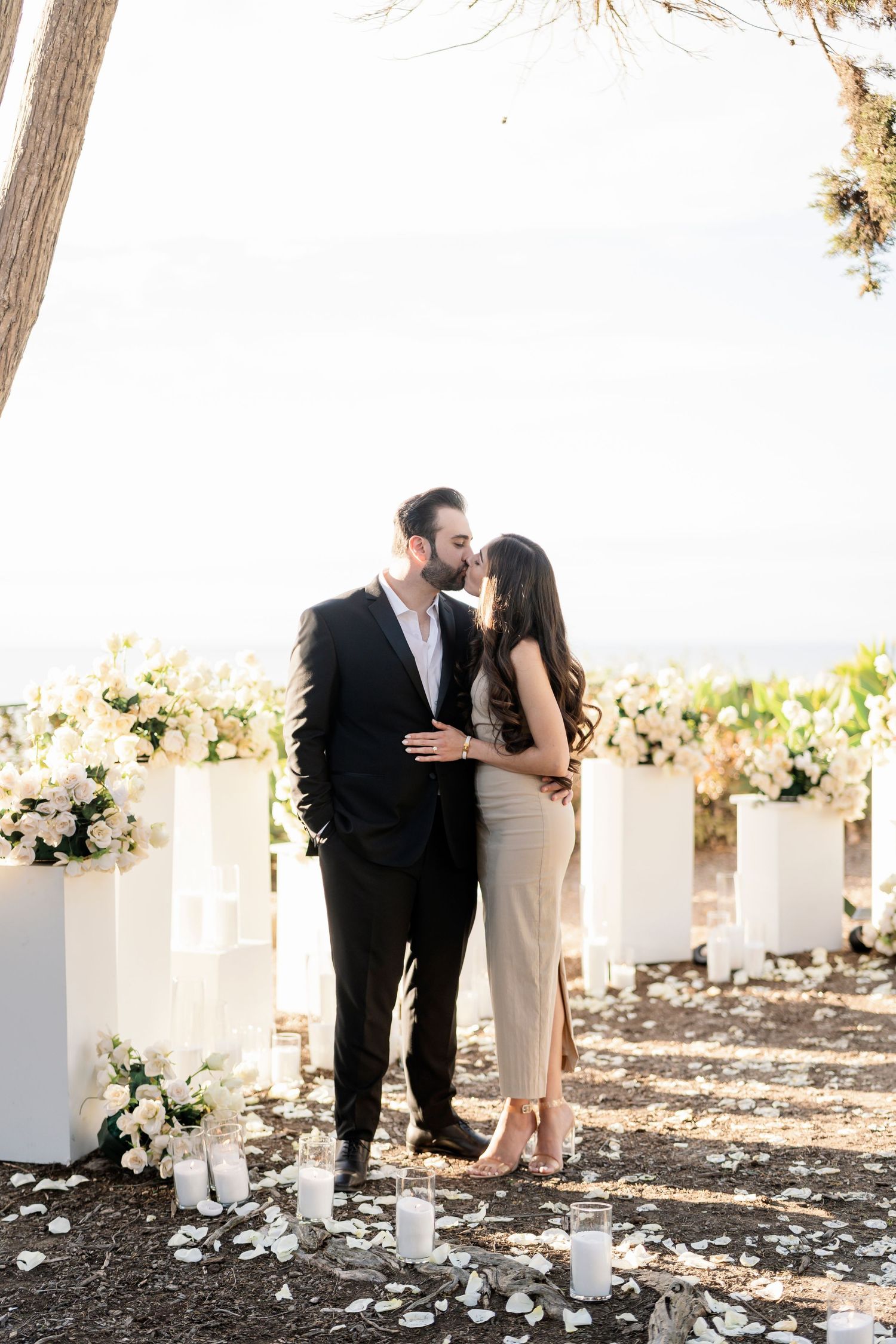 A couple shares a tender moment among white floral pillars and trees.