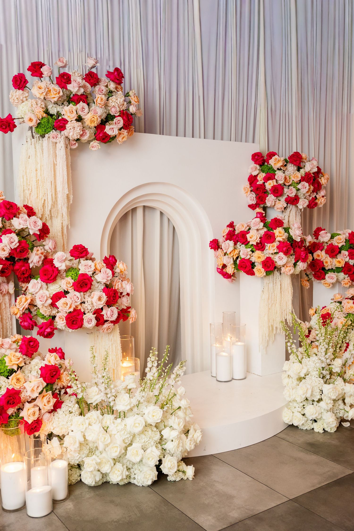 Elegant wedding backdrop with red and pink roses, white arch, candles, and floral arrangements for a romantic ceremony.