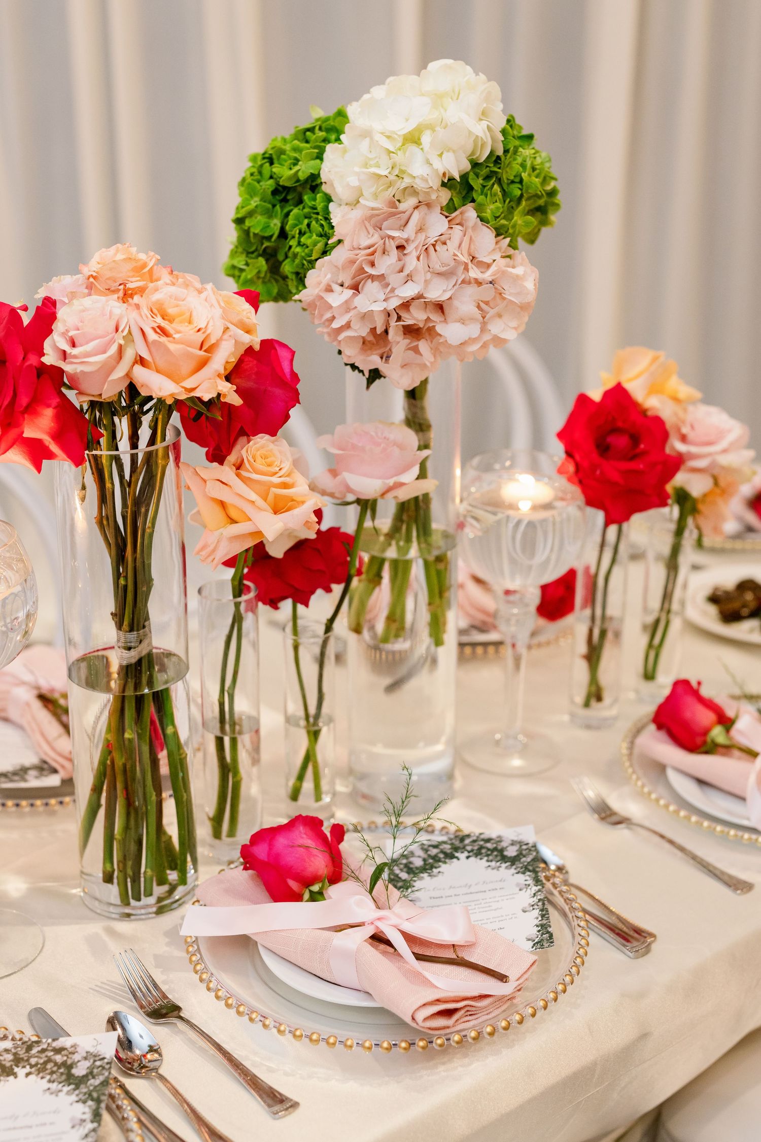 Elegant wedding table setting with pink hydrangeas, red and peach roses in glass vases, pearl-rimmed plates, and pink napkins.