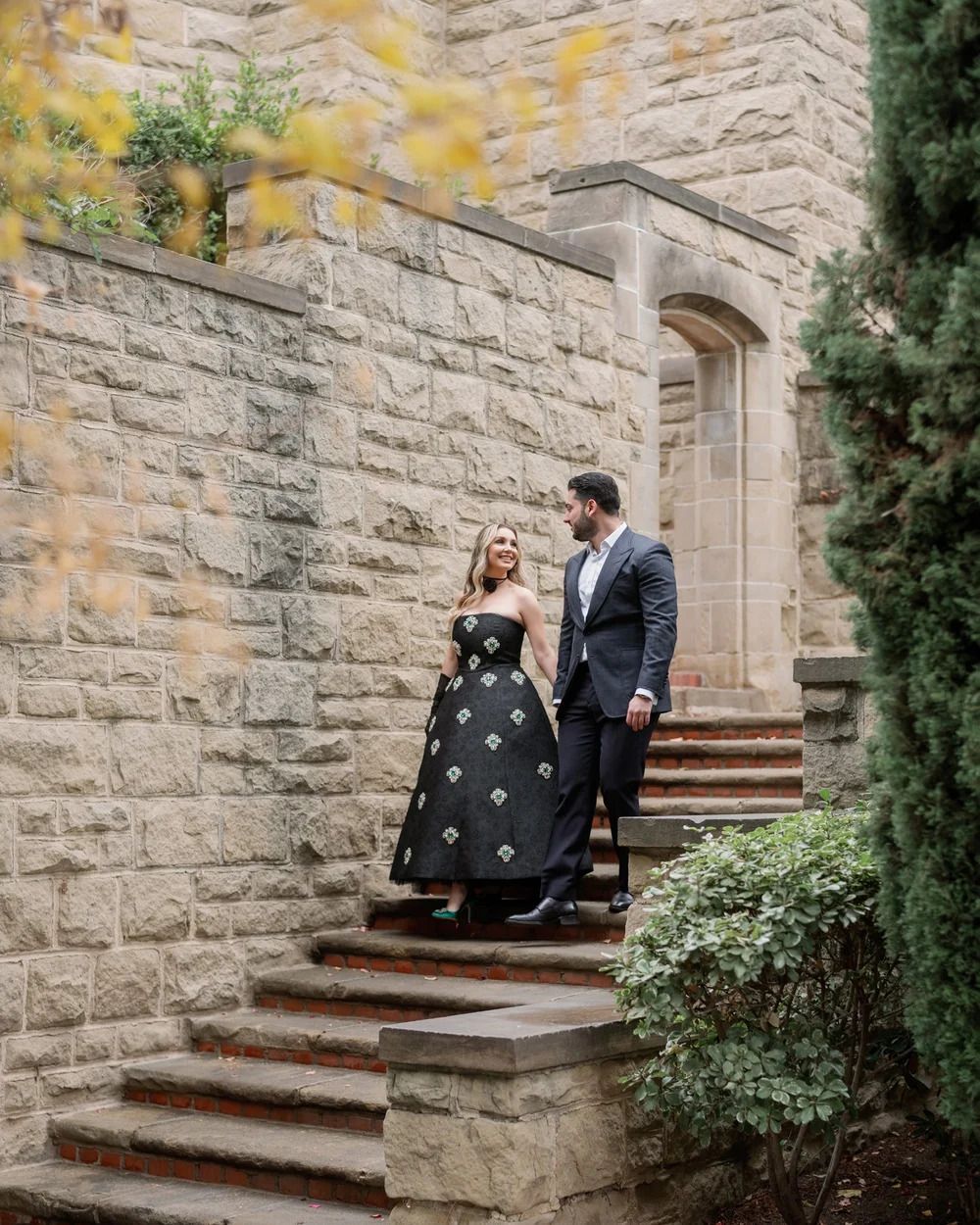 A couple descends elegant stone steps beside a historic building with autumn foliage.