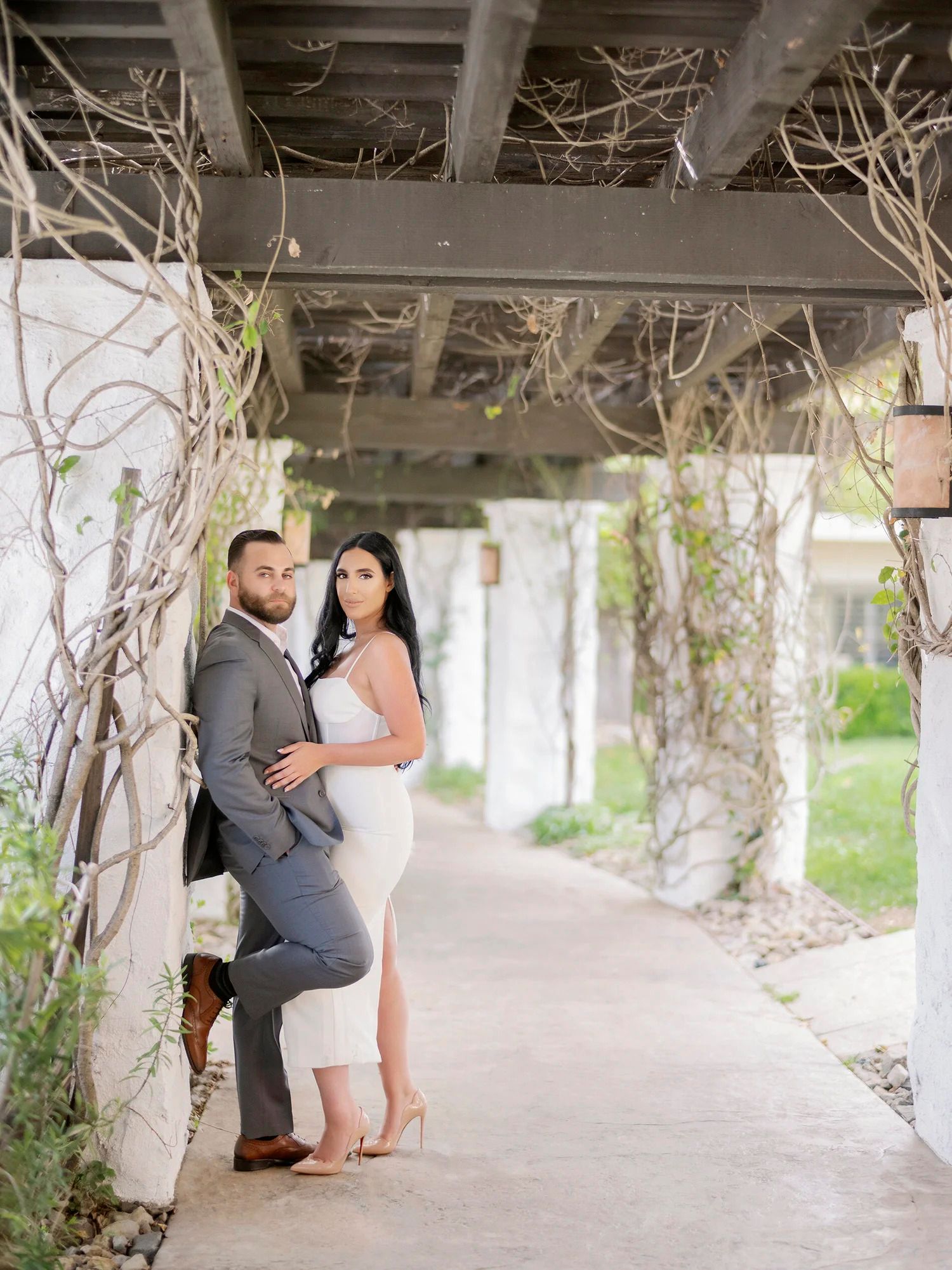 A couple embraces under a covered walkway with vine-wrapped columns.