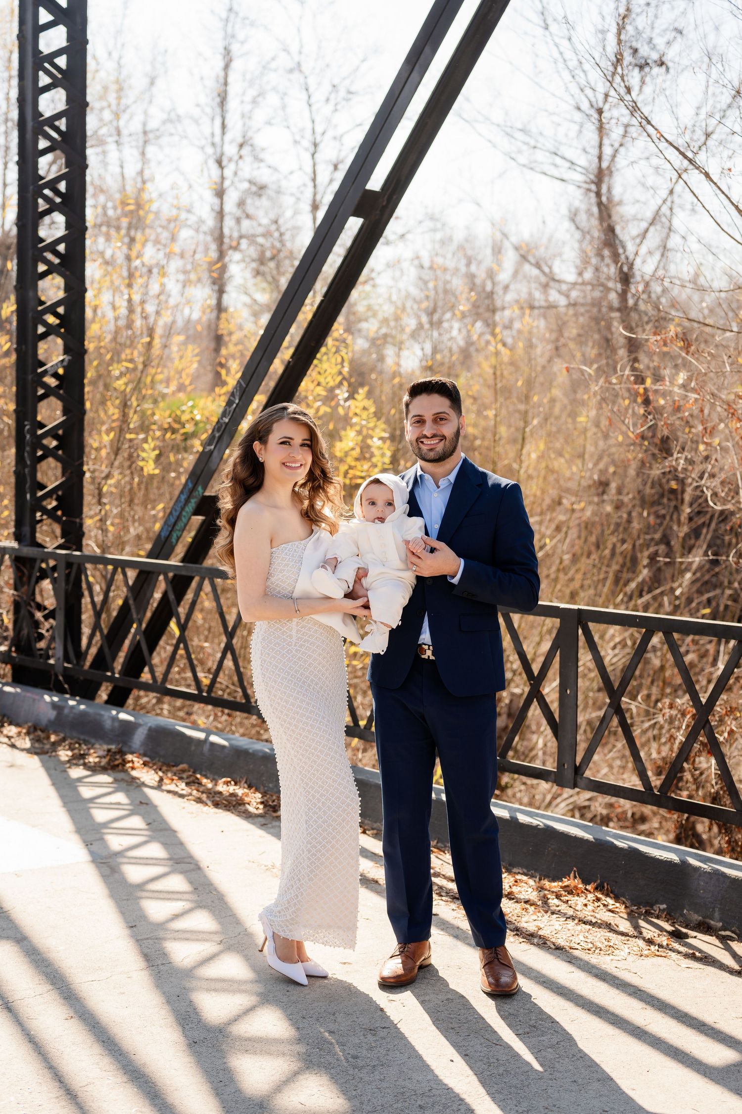 A couple in formal attire stands on a rustic bridge during a fall day with bare trees in the background.