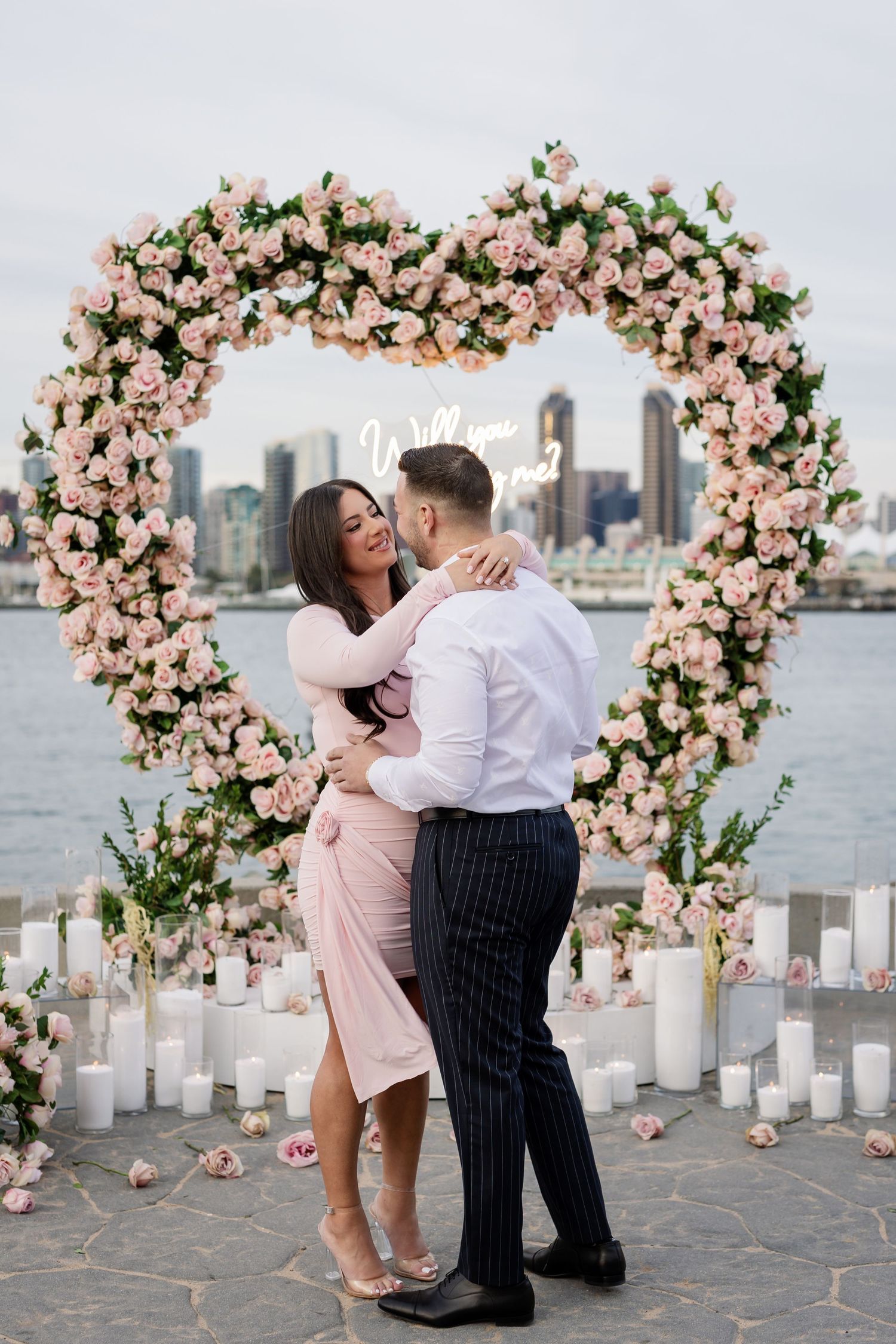 Couple celebrates special moment under pink floral arch with city skyline in background.