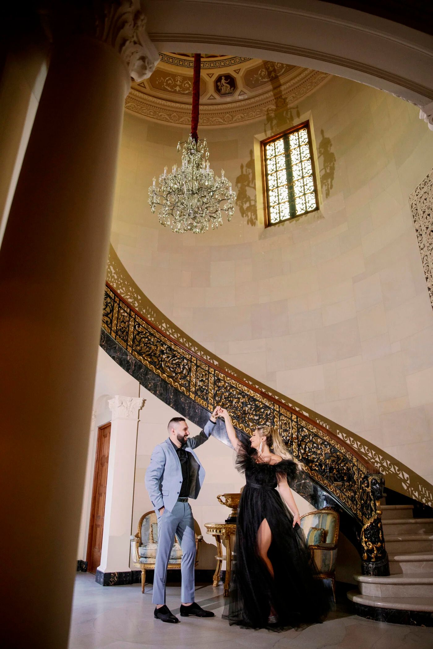 A couple dances beneath a crystal chandelier on an ornate spiral staircase in a grand interior.