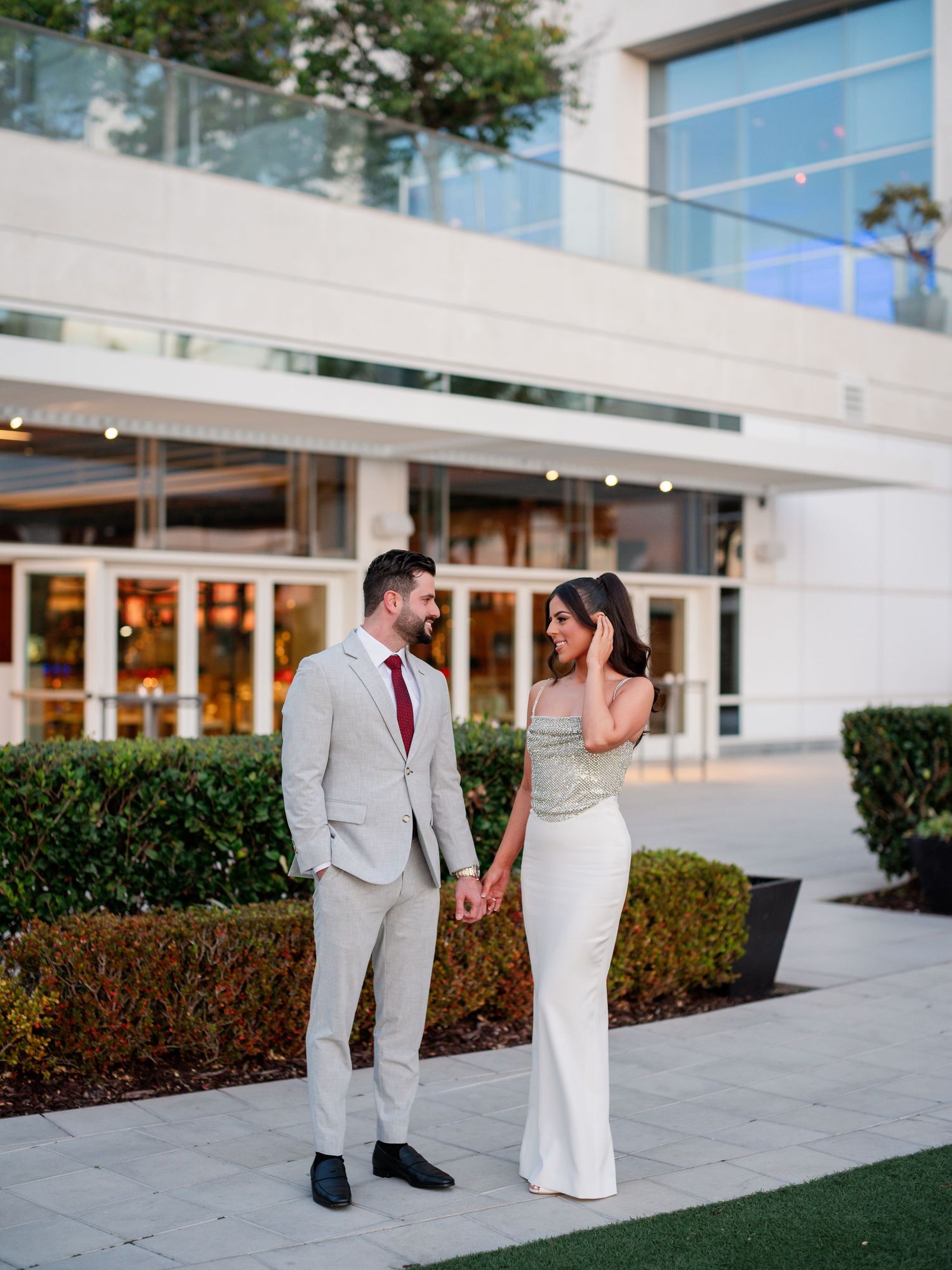 A couple poses outside a modern building with glass windows and grey exterior.