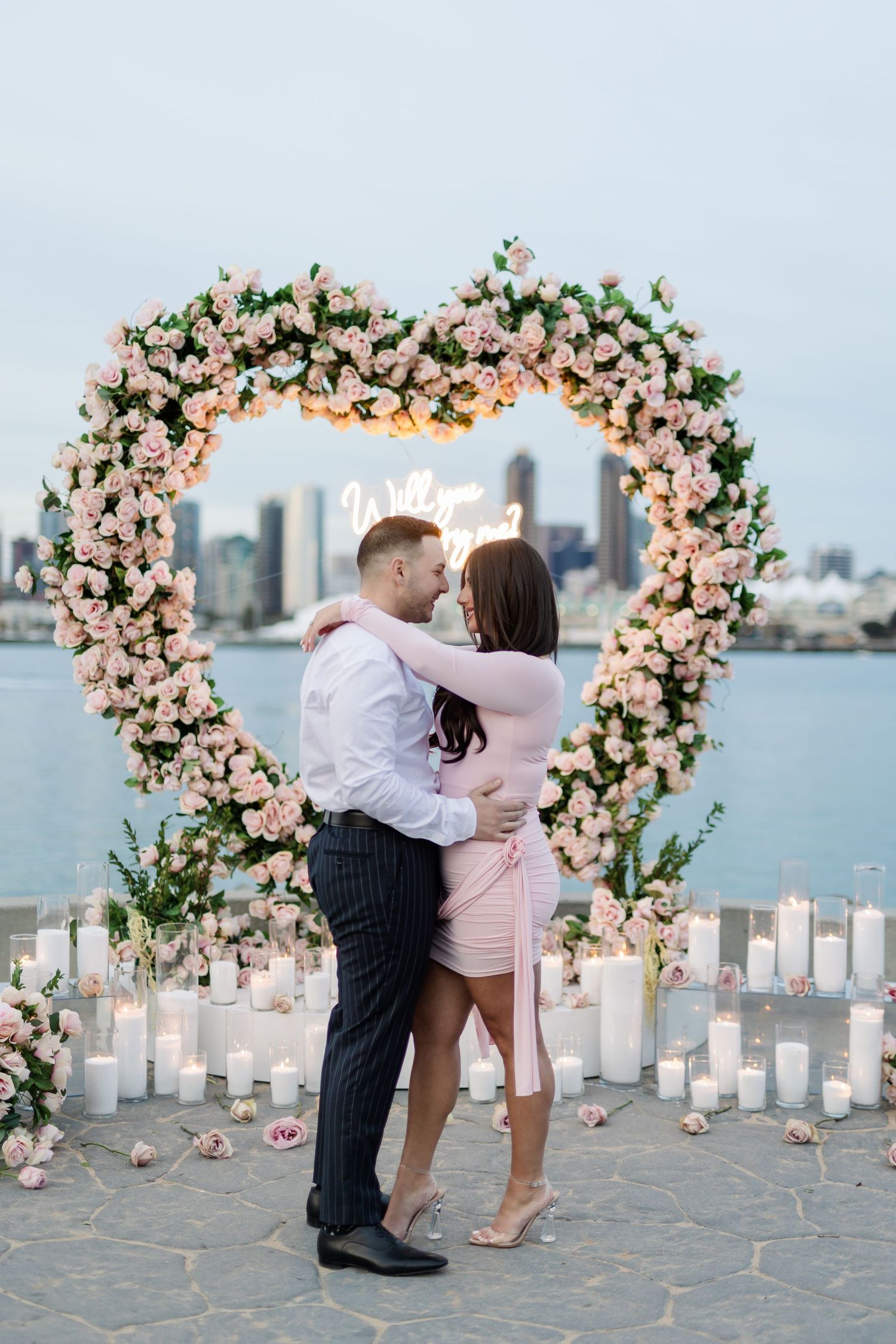 Couple sharing romantic moment under floral arch with candlelit pathway and city views.