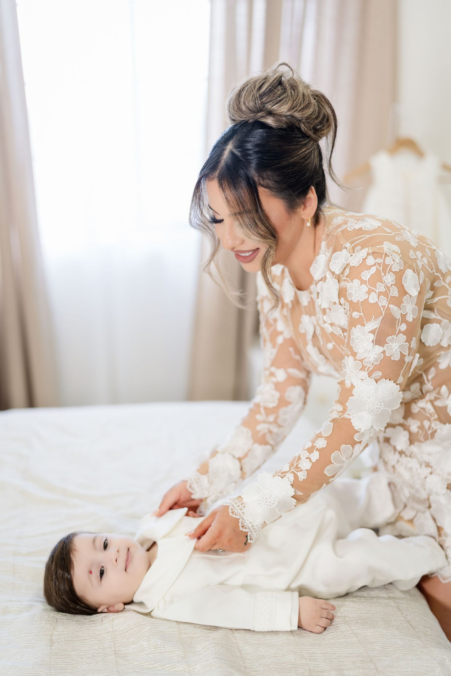 Mother in lace dress lying on bed playing tenderly with baby in white outfit on light bedding.