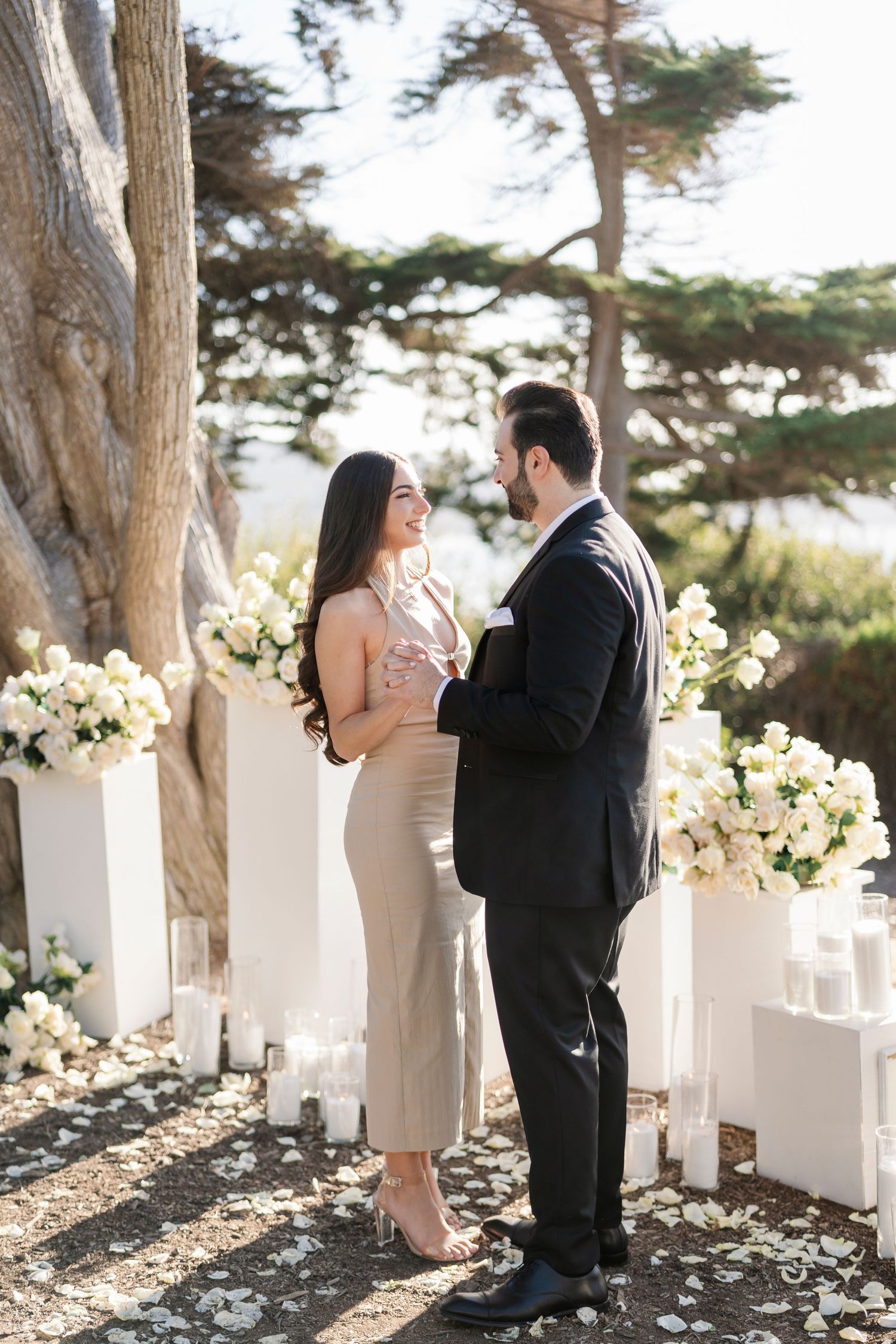 A romantic proposal scene unfolds between trees with white floral decorations.