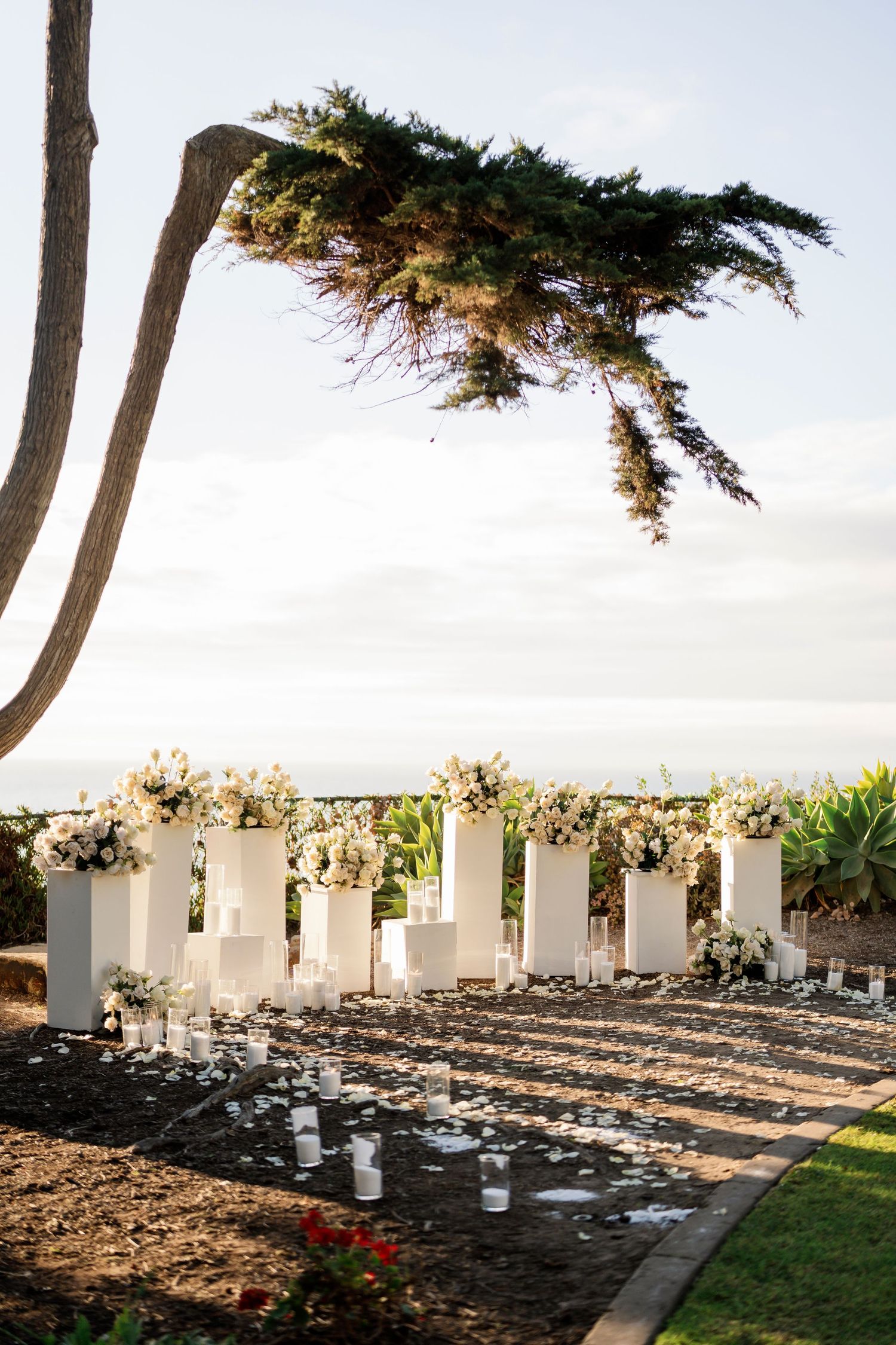 White floral arrangements line decorative pillars in outdoor garden setting with ocean view.
