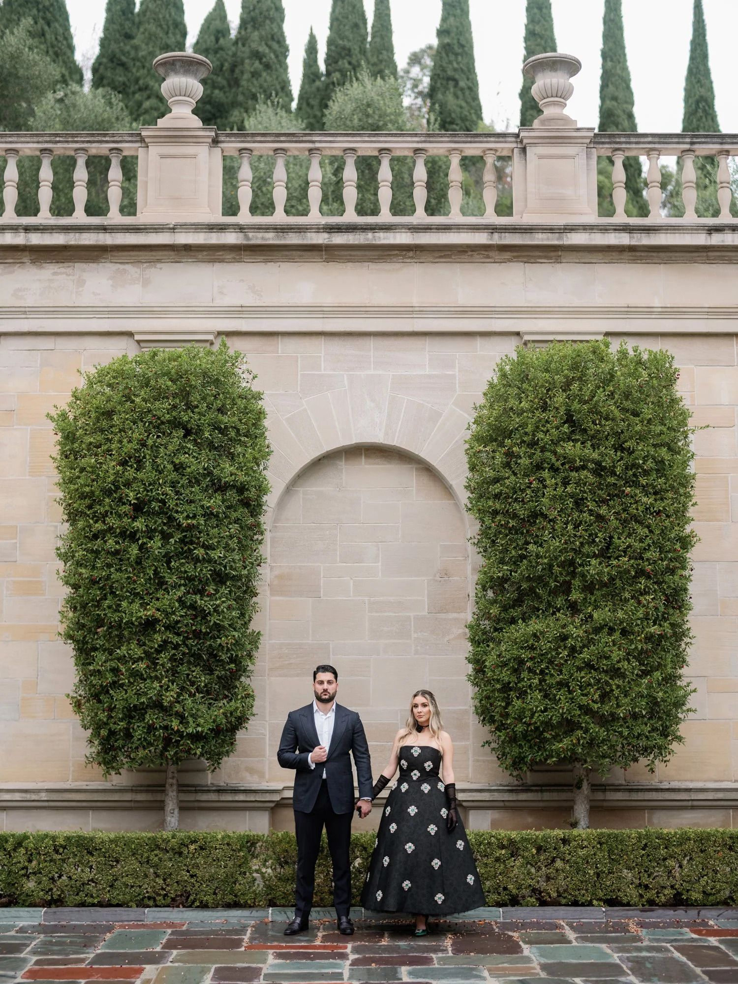 A couple poses in formal attire against a stone wall with manicured hedges and classical architecture.