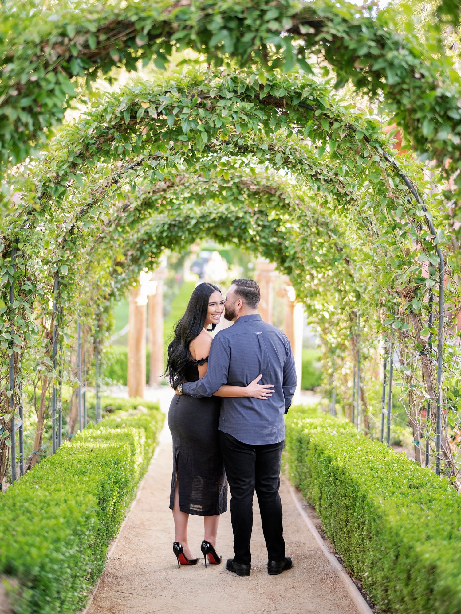 A couple walks through a romantic garden tunnel covered in climbing vines and greenery.
