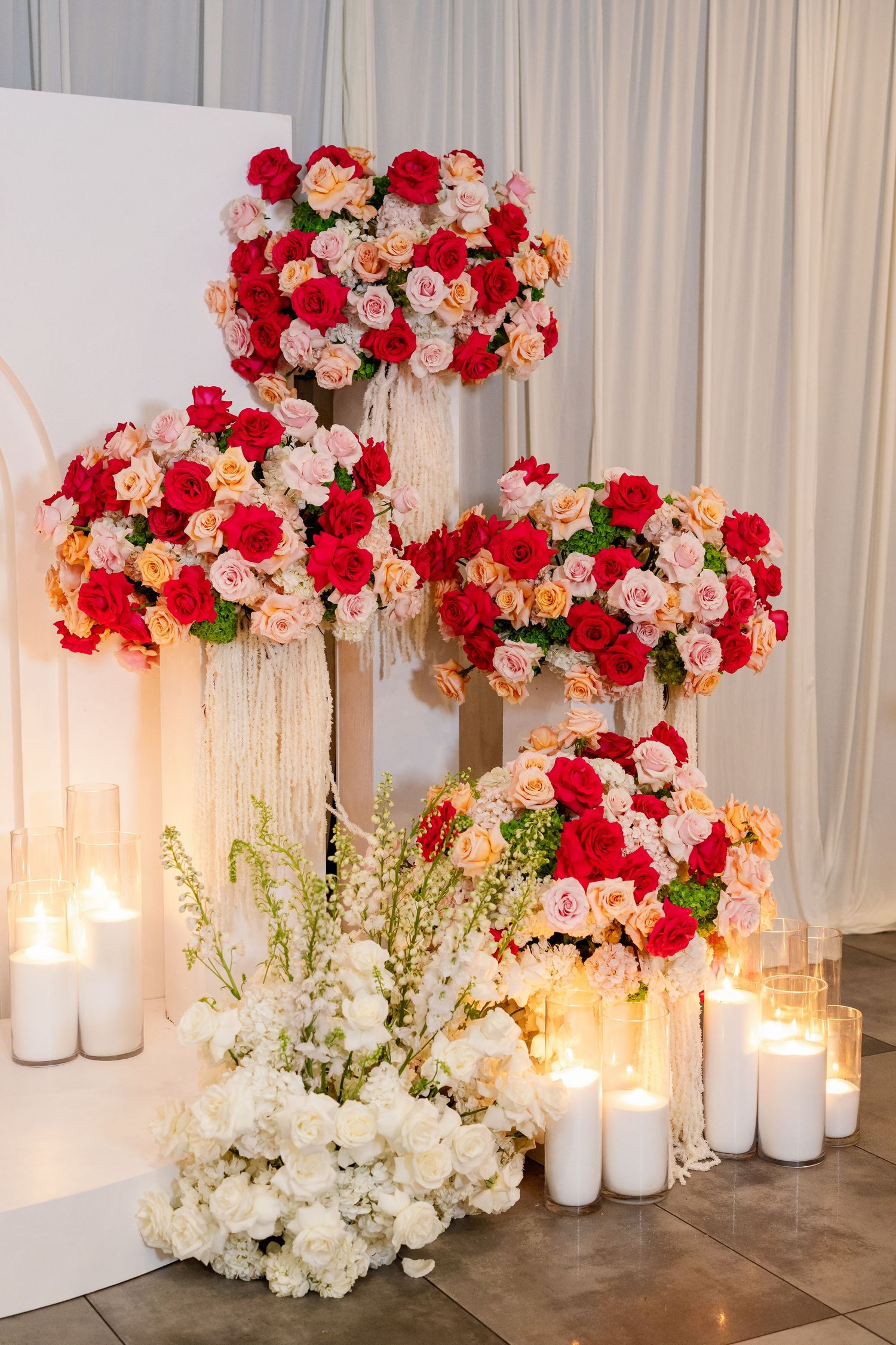 Elegant wedding floral display with red, pink roses on tall stands, white candles, and white flower arrangements on floor.