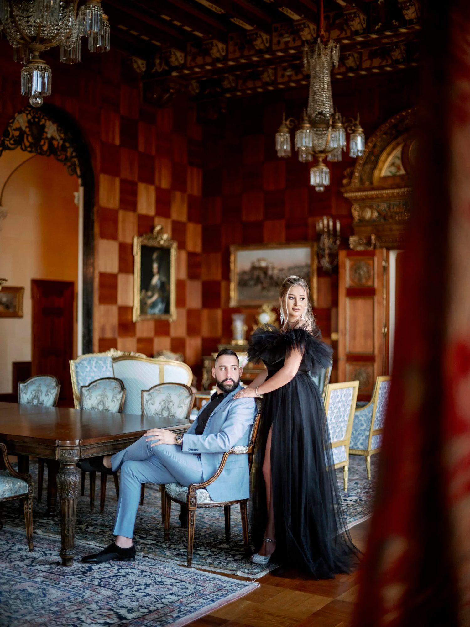 A couple poses in an elegant room with checkerboard walls and vintage furniture.