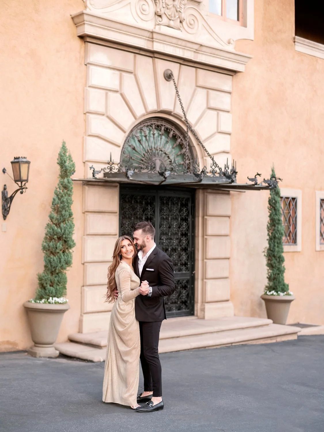 Romantic scene against peach-colored building exterior with ornate architectural details and potted cypress trees.
