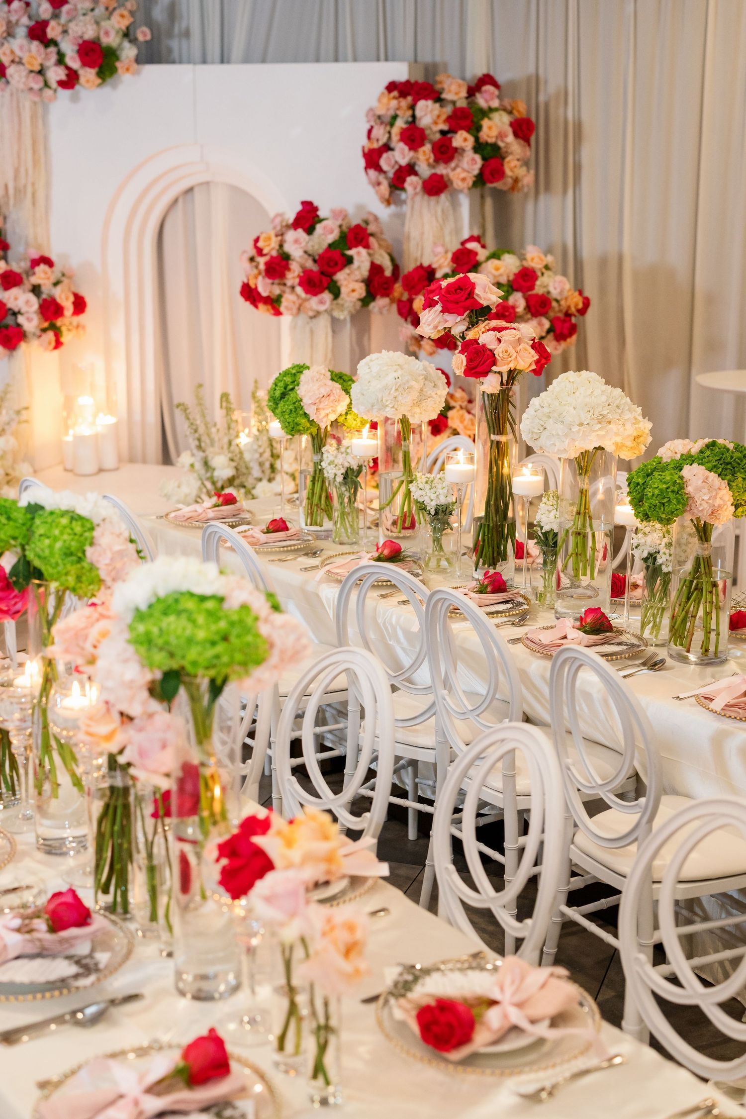 Elegant wedding reception table with white chairs, red and pink floral centerpieces, candles, and fine dining place settings.