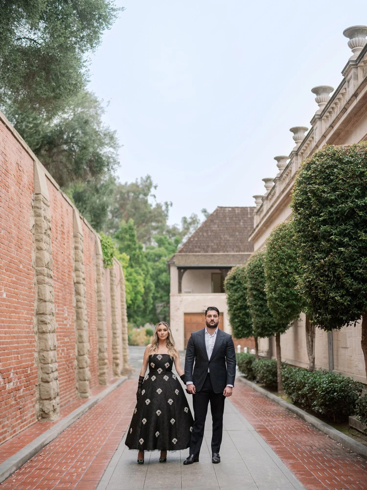 A couple walks along a brick-lined path between historic walls and manicured hedges.