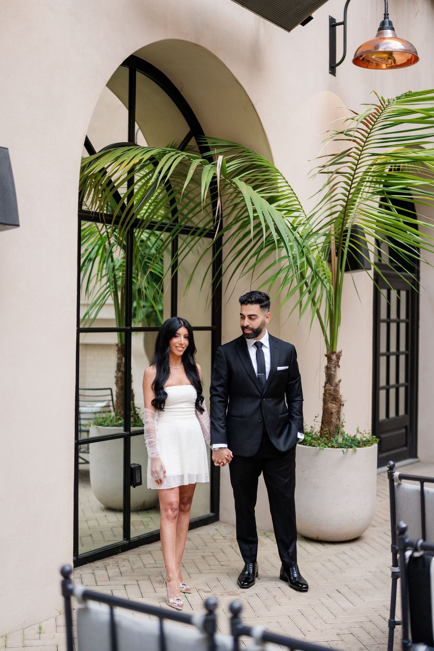 Couple in formal wear walking together along Mediterranean-style courtyard with palm trees.