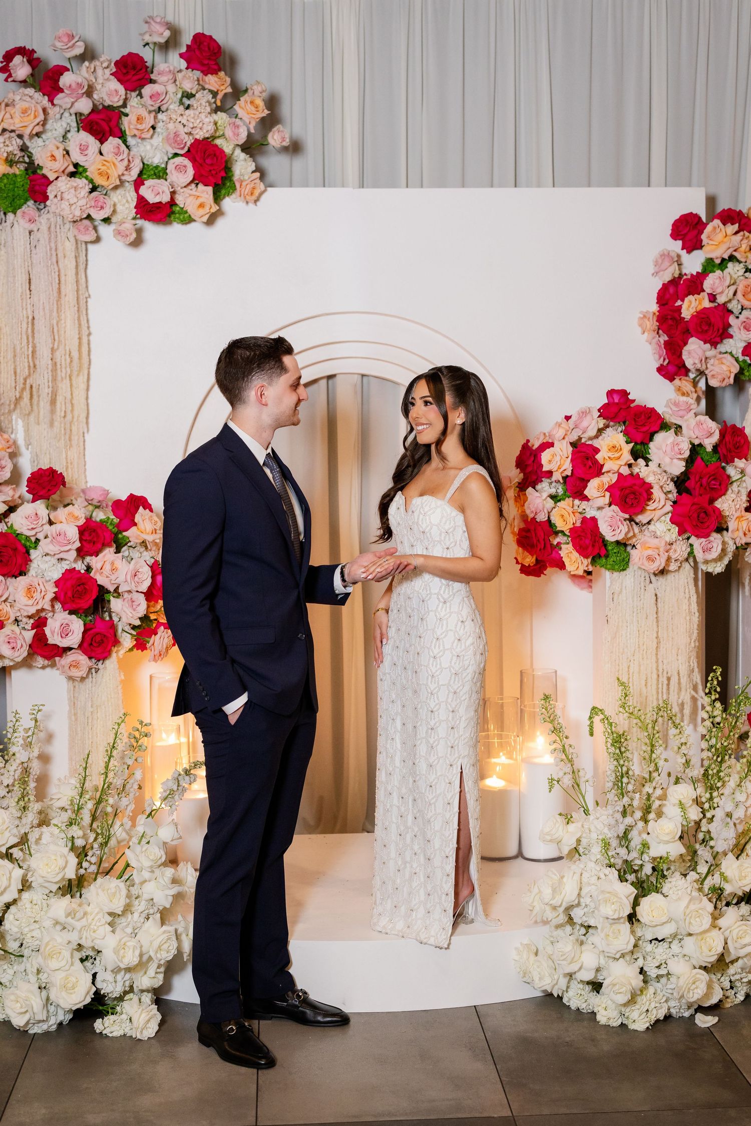 Couple exchanging vows at floral altar with red and pink roses, white blooms, and candles in elegant wedding ceremony.