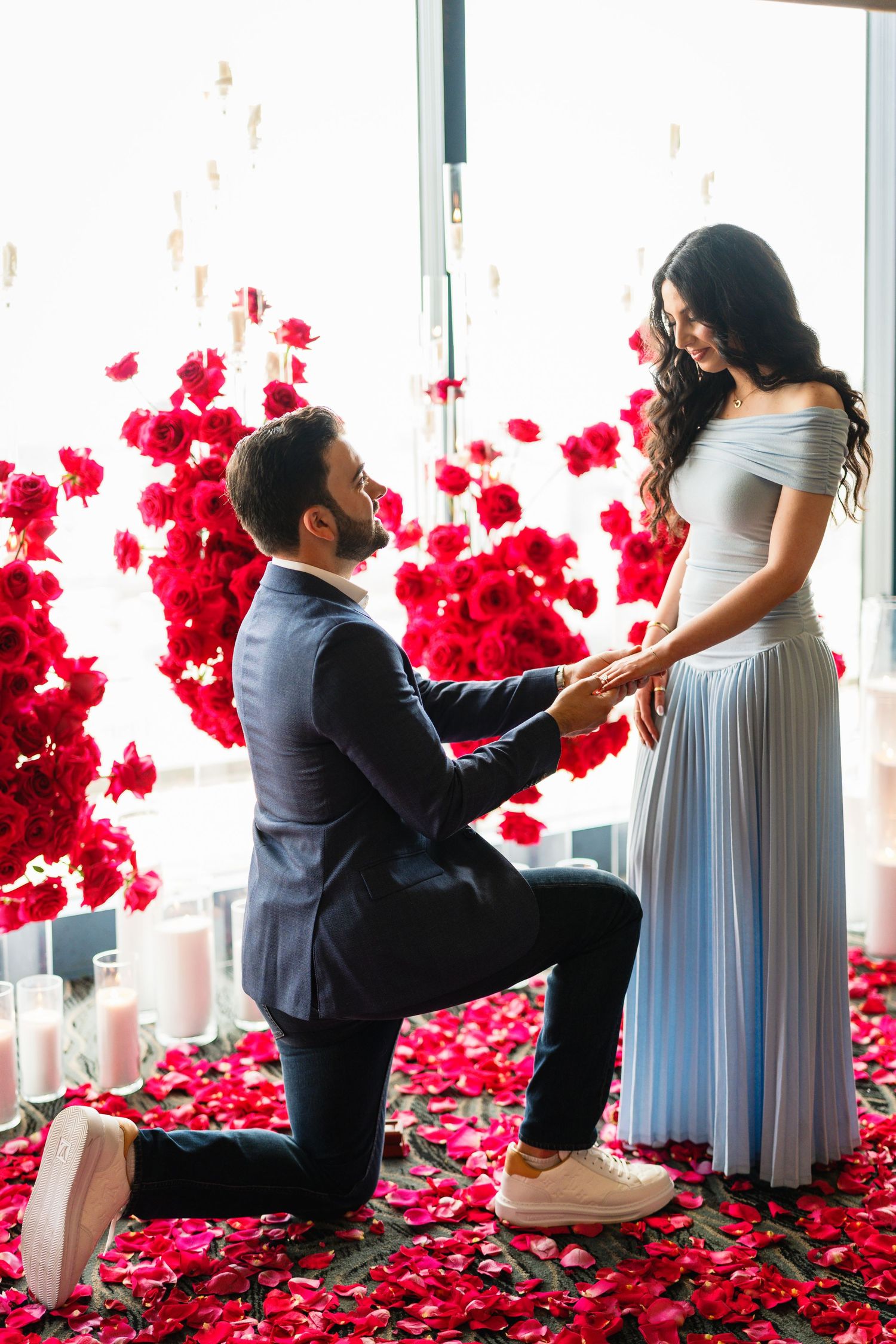 A proposal moment captured with red rose petals and city views through large windows.