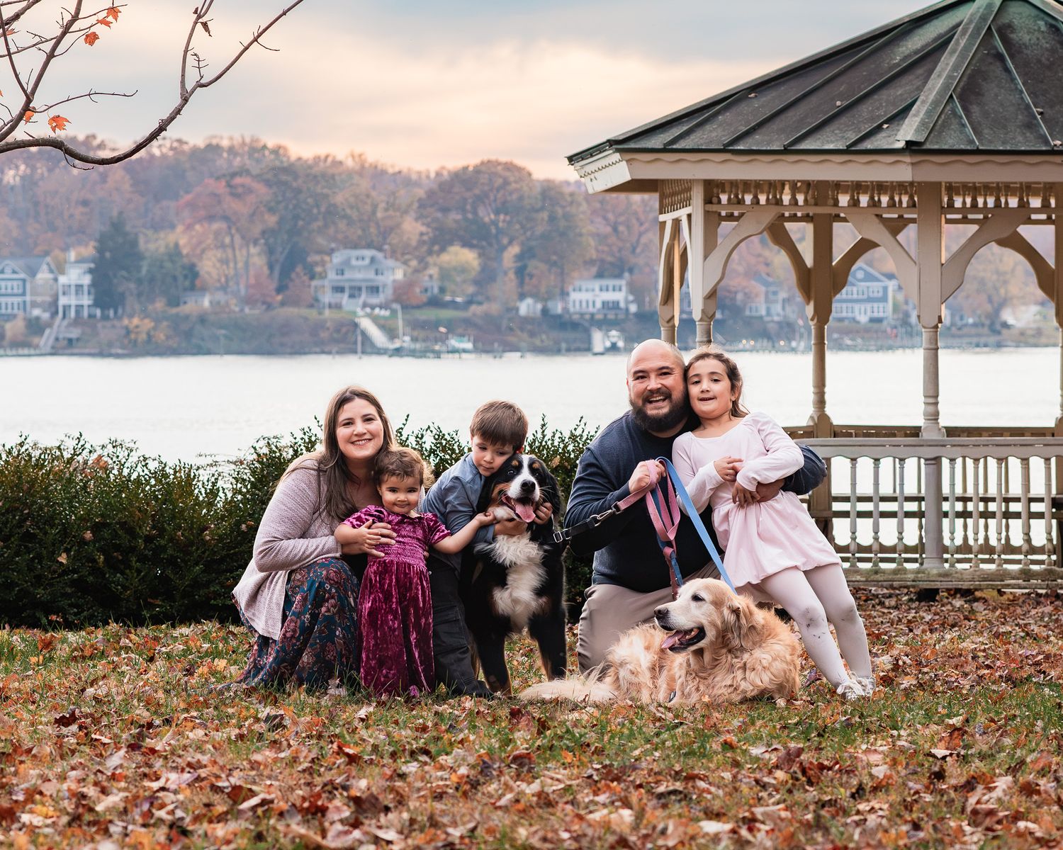 Family of five with two dogs posing for a fall family portrait at the waterfront in an Annapolis Park next to a gazebo.