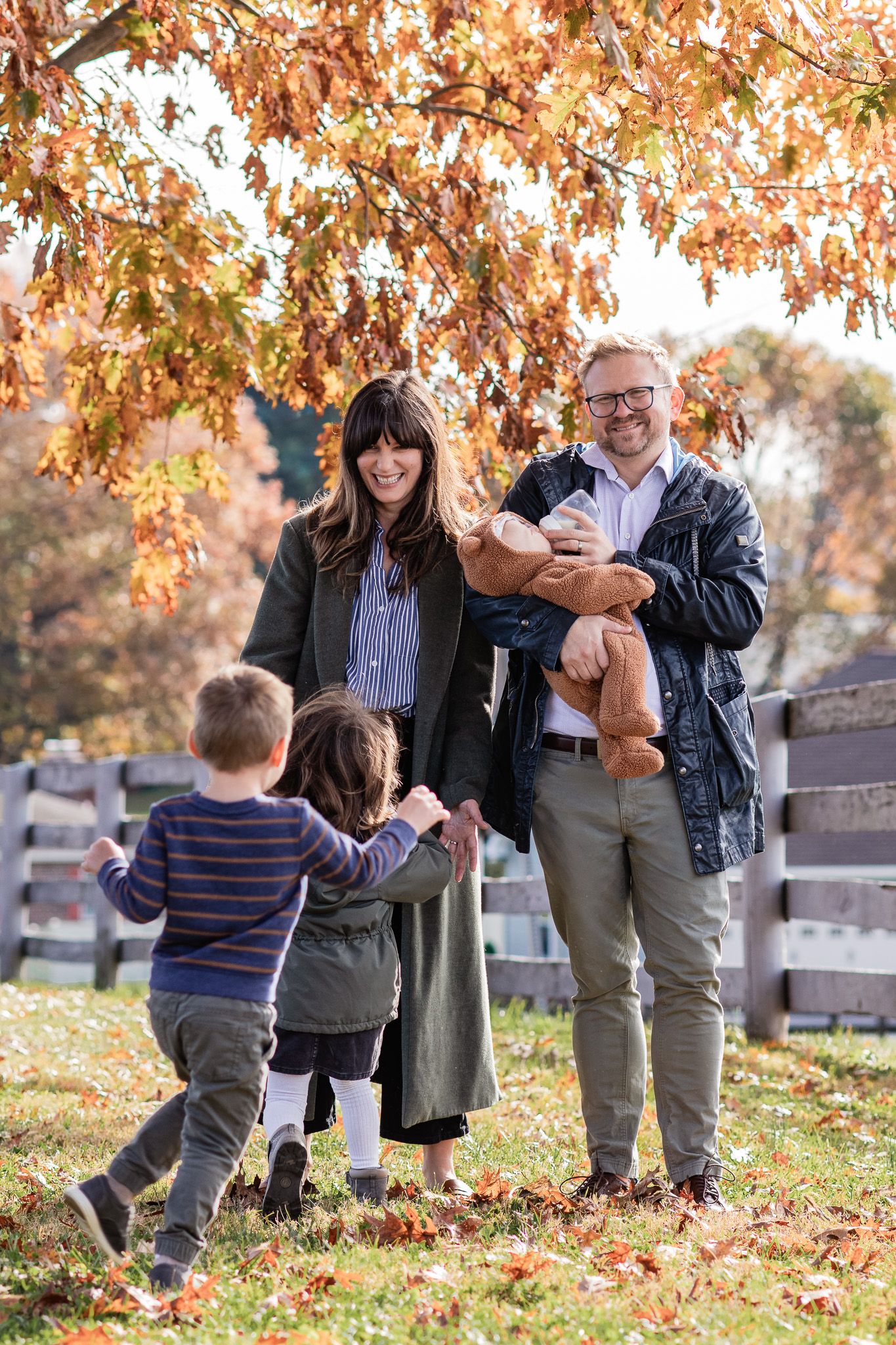 Family of five standing in front of a fence and tree with autumn leaves, kids running to their parents holding the baby.