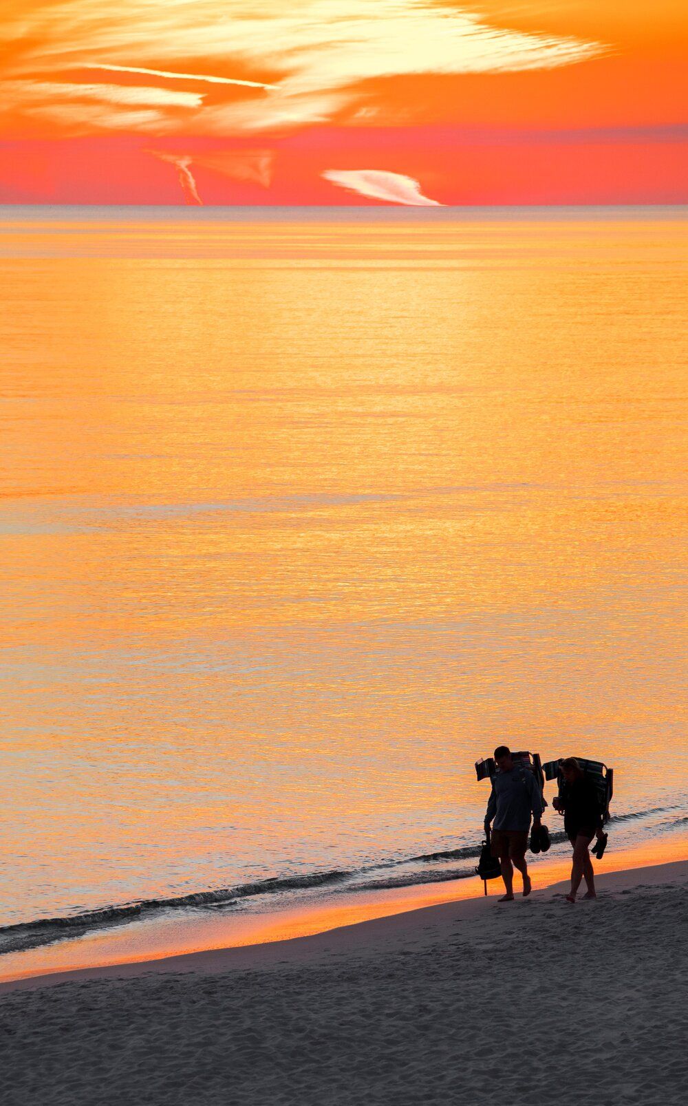 Silhouettes of people walking along a beach at sunset with vibrant orange sky reflecting on calm ocean waters.