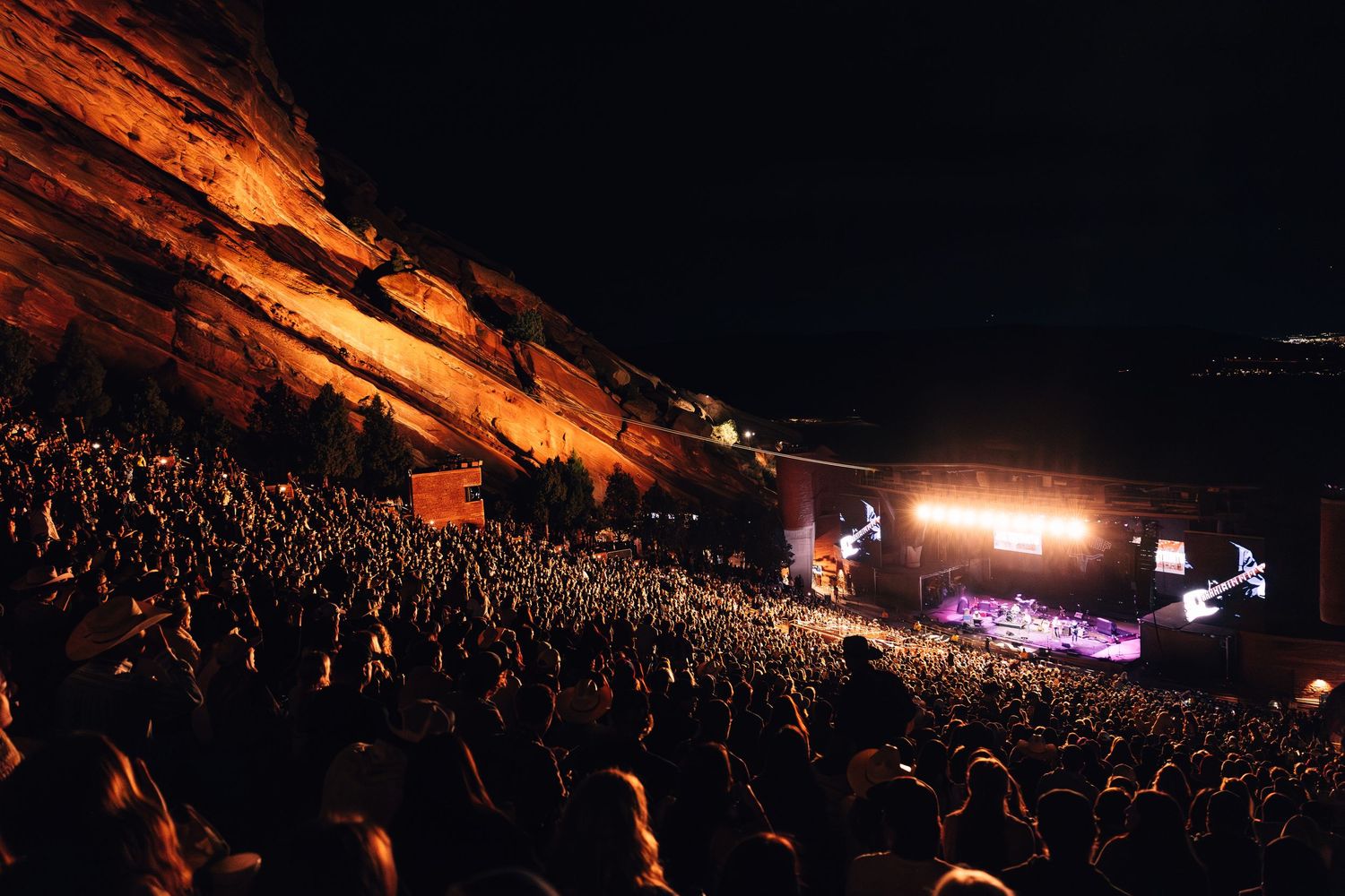 Concert crowd silhouettes against illuminated stage at Red Rocks Amphitheatre during nighttime performance.