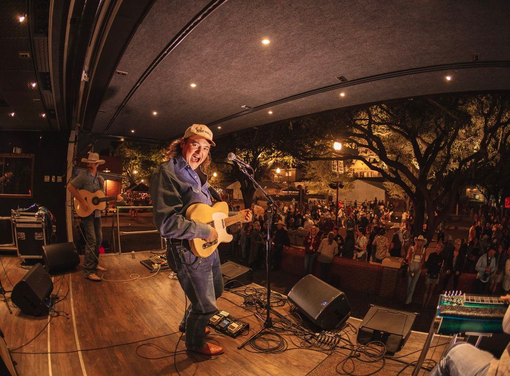 Guitar player performs on outdoor stage at night with crowd in background.