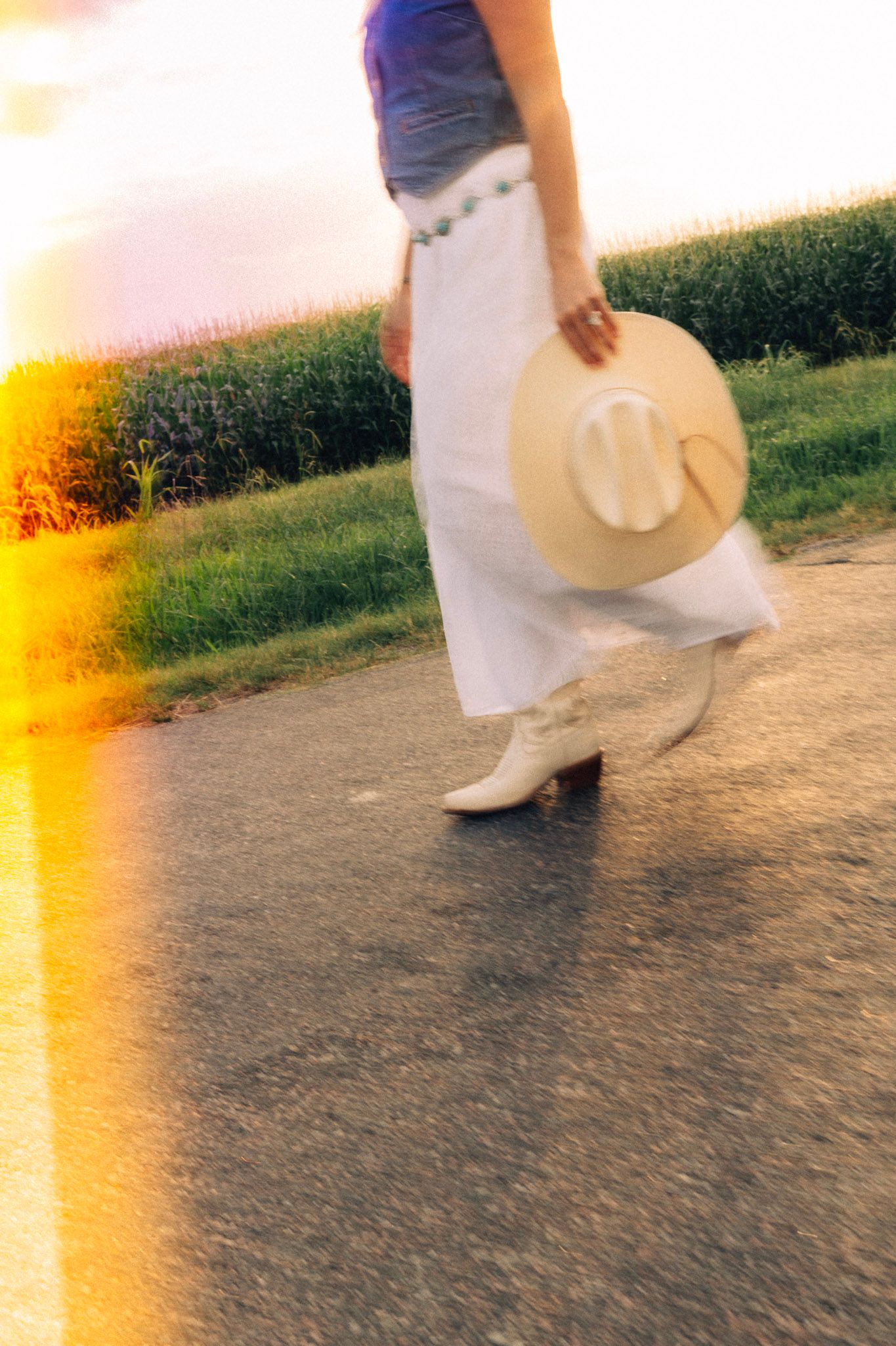 Woman in white skirt and boots holding a sun hat walks along a sunlit path at golden hour.