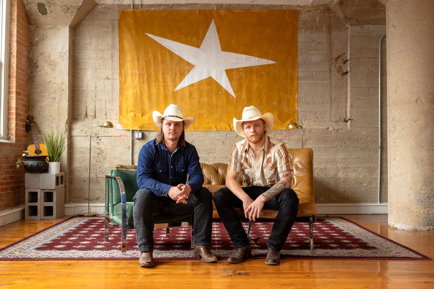 Two men in cowboy hats sit on a couch before a large star flag in a rustic loft space.