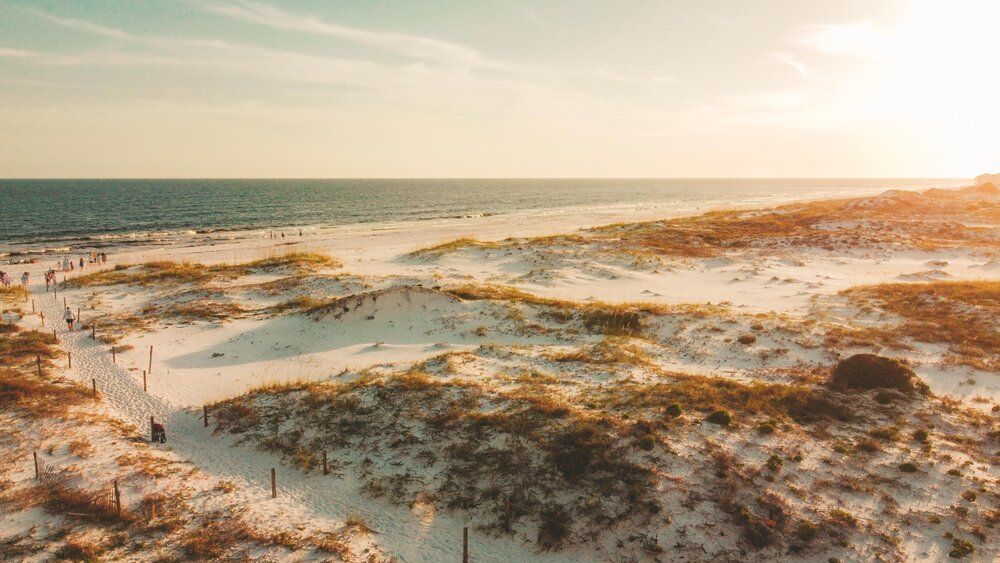 Aerial view of sandy beach dunes at sunset with wooden boardwalk path leading to ocean.