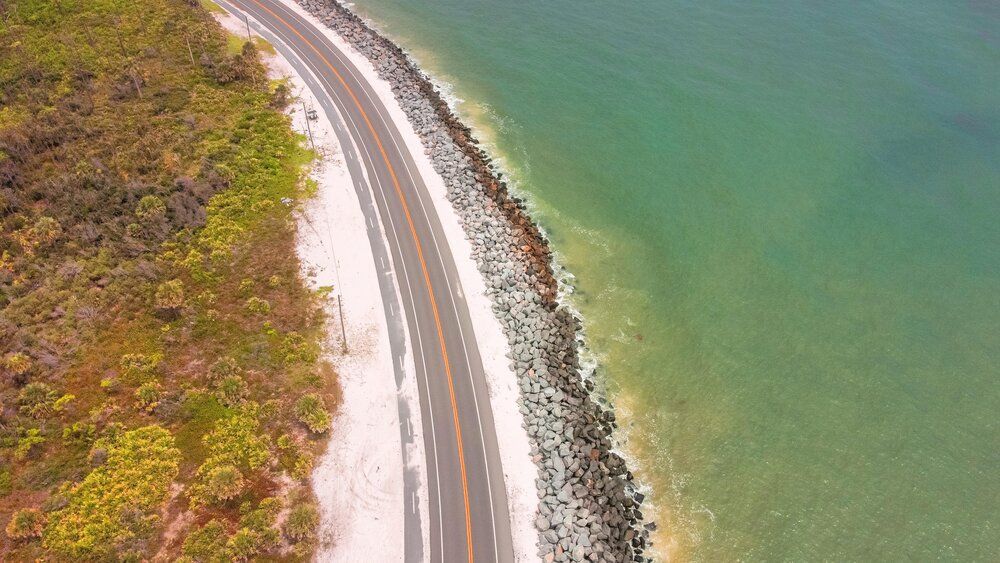 Aerial view of a coastal highway winding along turquoise waters with rock barriers and sandy beach.