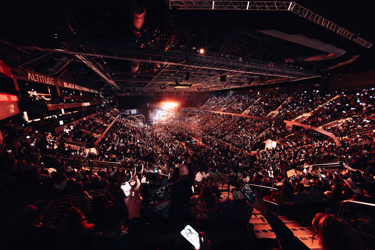 Large indoor arena filled with a massive crowd during a concert or event, illuminated by dramatic red lighting.