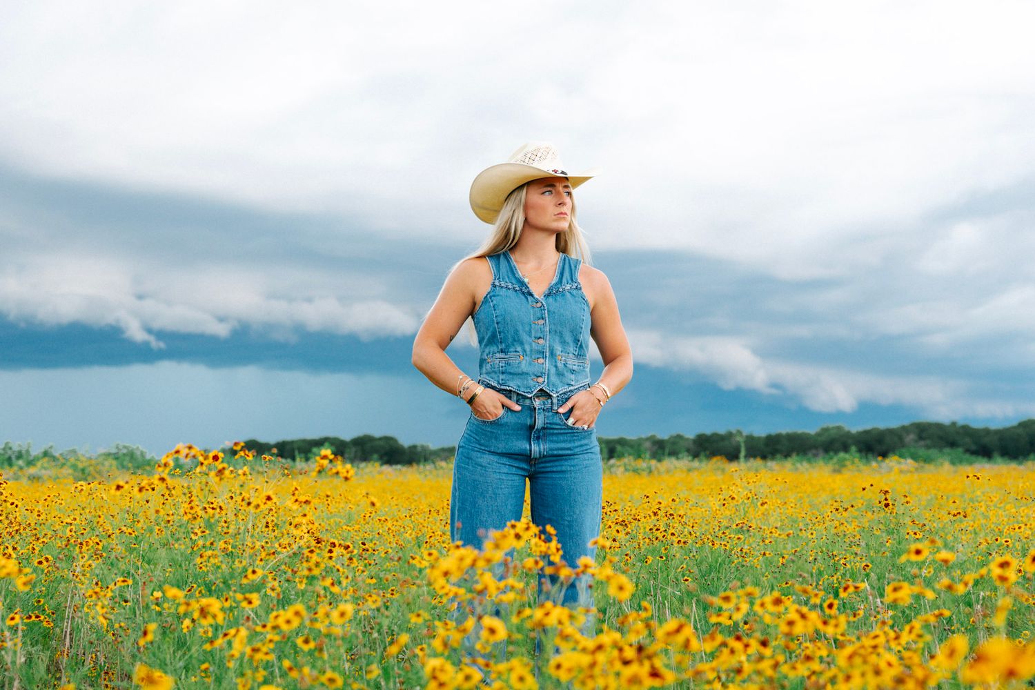 Woman in cowboy hat and denim overalls standing in yellow wildflower field under dramatic cloudy sky.
