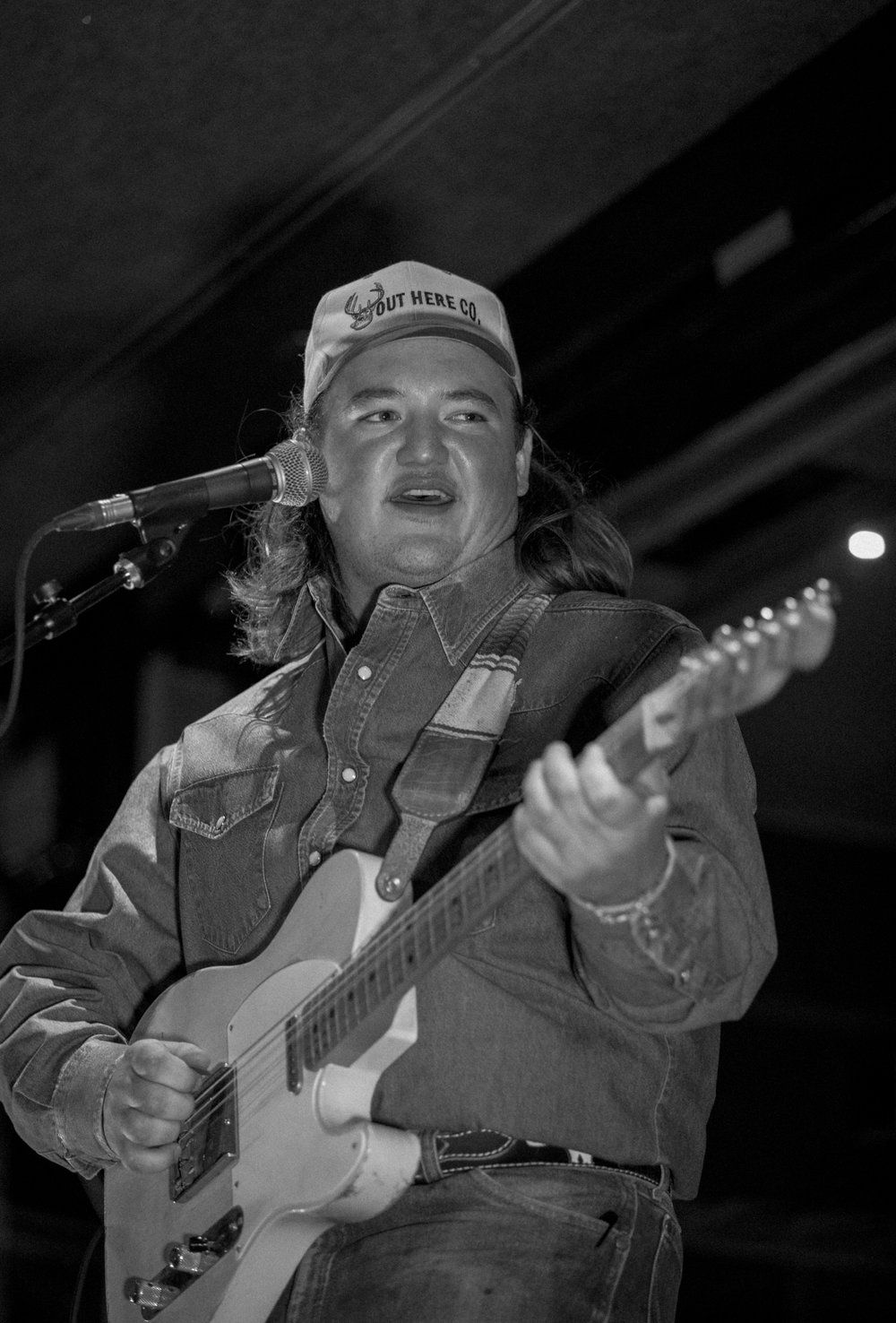 Performer plays electric guitar while singing into microphone wearing baseball cap and denim jacket.