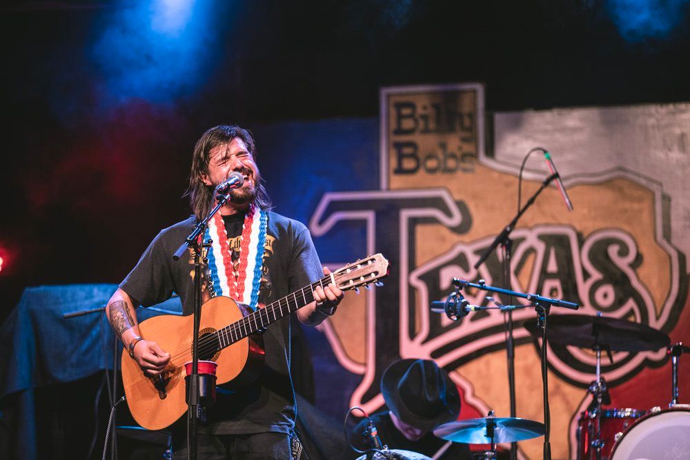 Musician performs with acoustic guitar on stage with Texas themed backdrop.