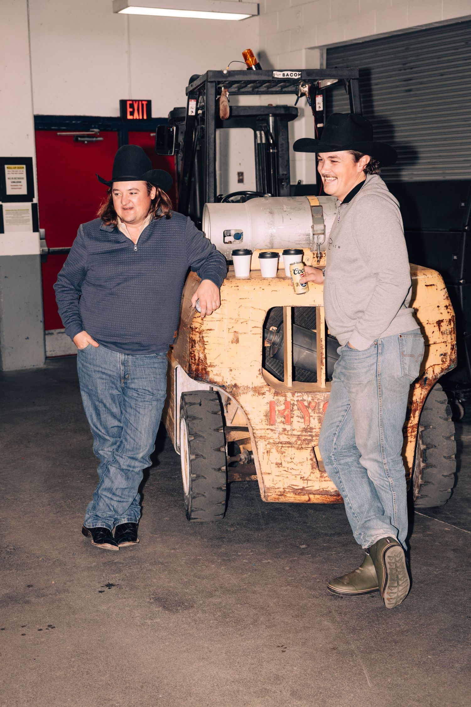 Two people in cowboy hats lean against a rustic wooden table with bottles in an industrial warehouse setting.
