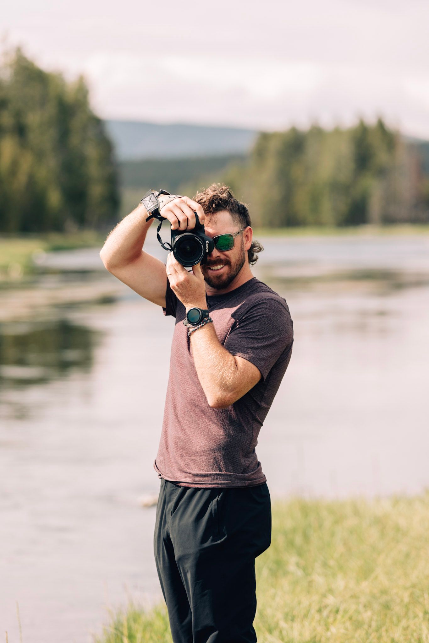 Photographer with a professional camera takes photos near a serene lake surrounded by forest and mountains.
