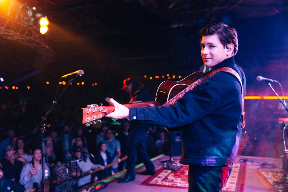 Performer in navy jacket plays guitar on dark stage with colorful lighting and crowd in background.