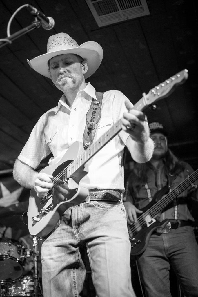 Country musician in white cowboy hat and jeans plays electric guitar on small stage.