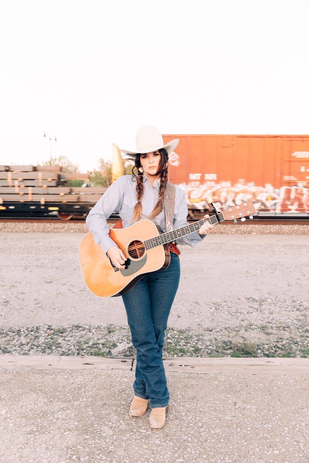 Country musician with an acoustic guitar poses against vintage train cars in casual western attire.