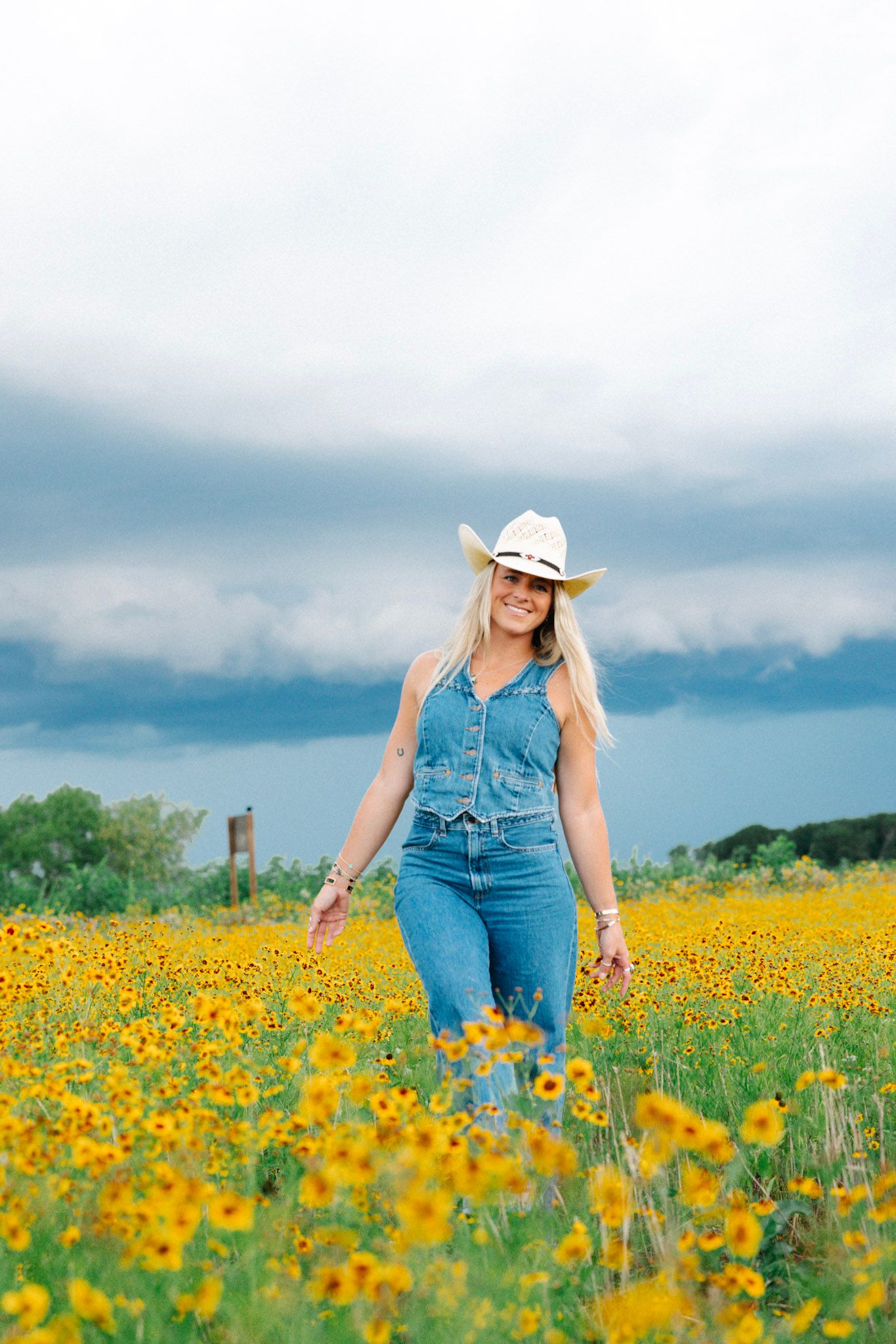 A smiling blonde woman in denim overalls and a white cowboy hat walks through a vibrant yellow wildflower field under cloudy skies.