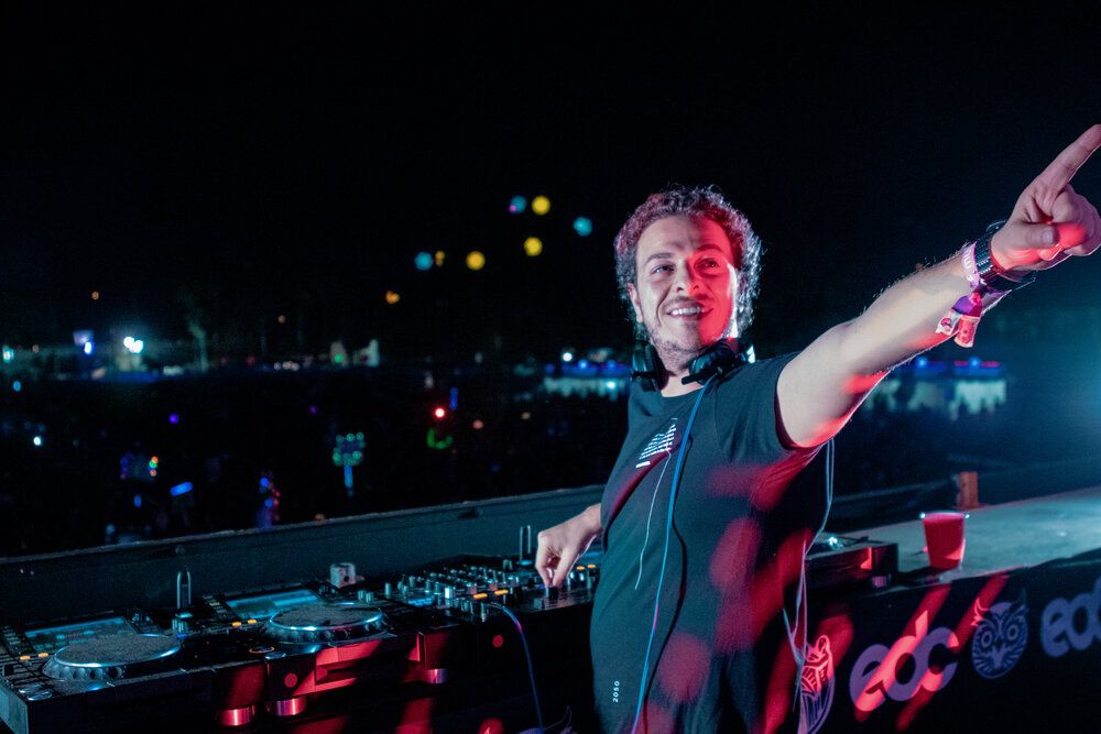 DJ performing at night on stage with red lighting and raised arm pointing to crowd at outdoor music festival.