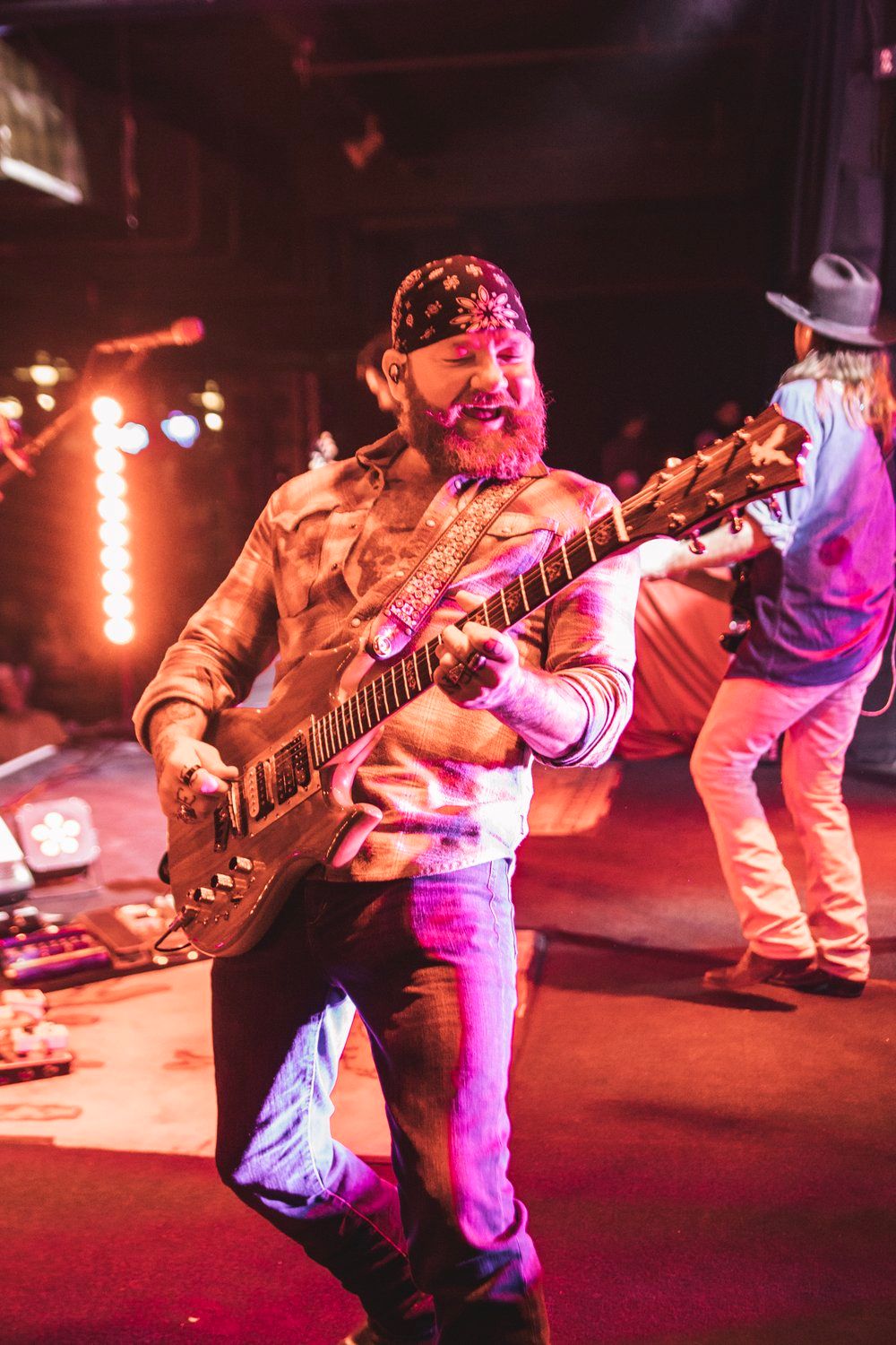 Guitarist in bandana performs energetically on stage with red concert lighting.