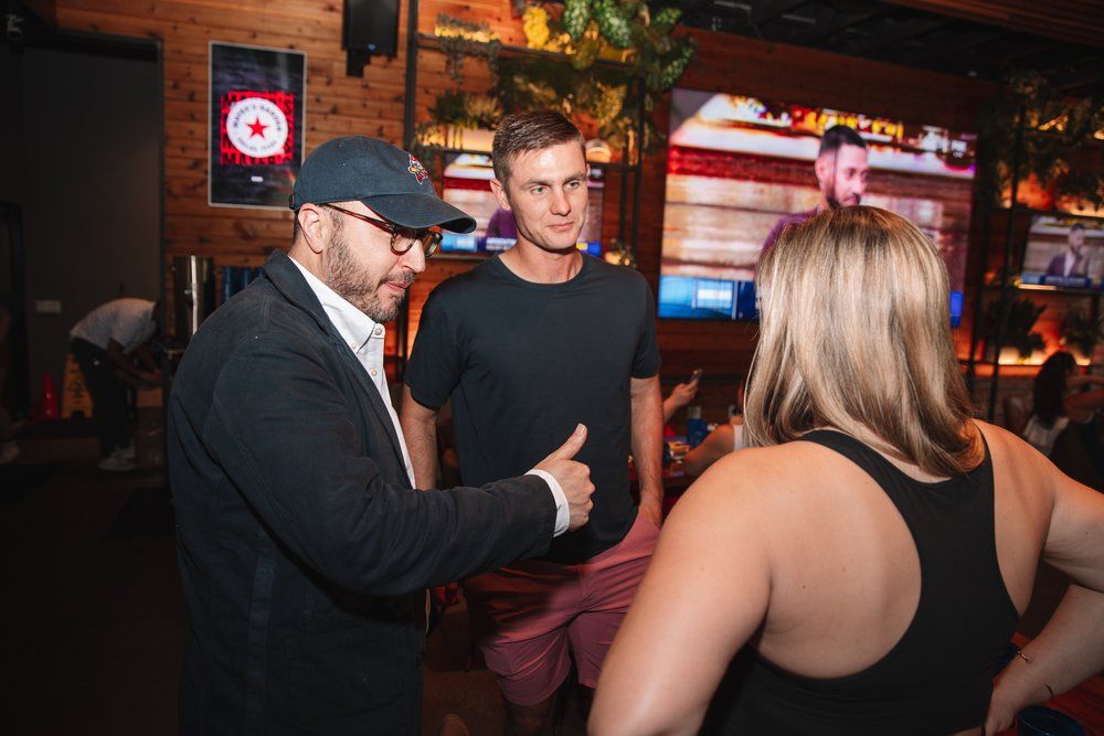 Group engaged in animated conversation at a dimly lit bar with neon signs visible in the background.
