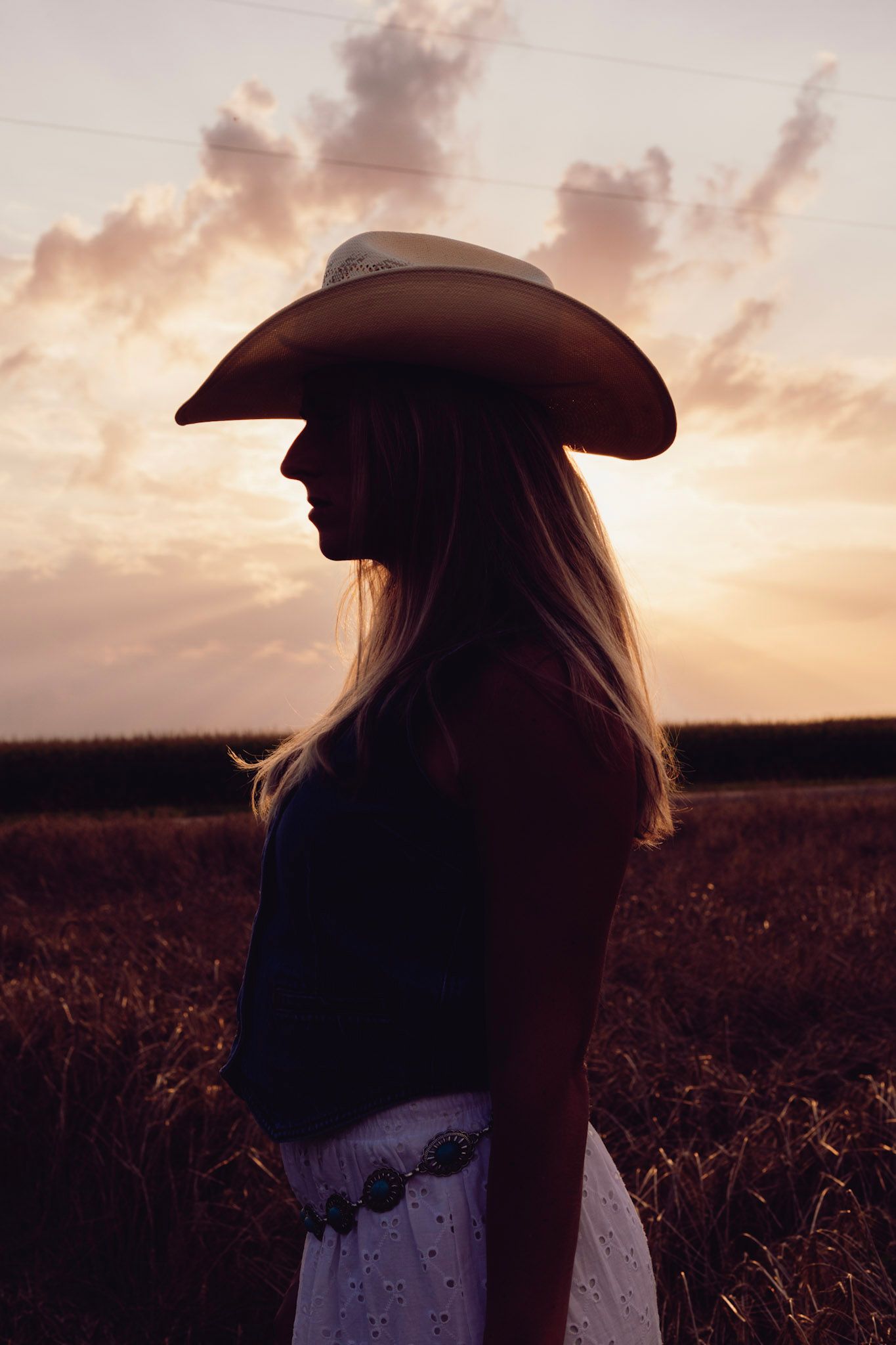 Silhouette figure wearing a cowboy hat and dress stands in a field during golden sunset with dramatic clouds.