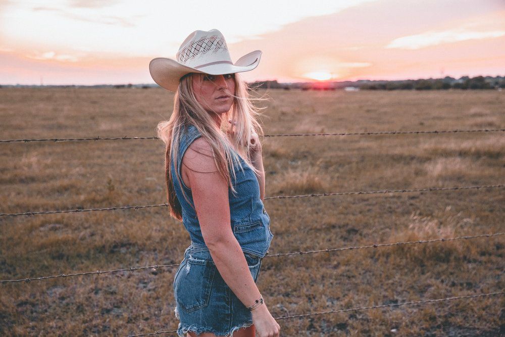 A person in denim overalls and cowboy hat stands in a golden field during sunset.